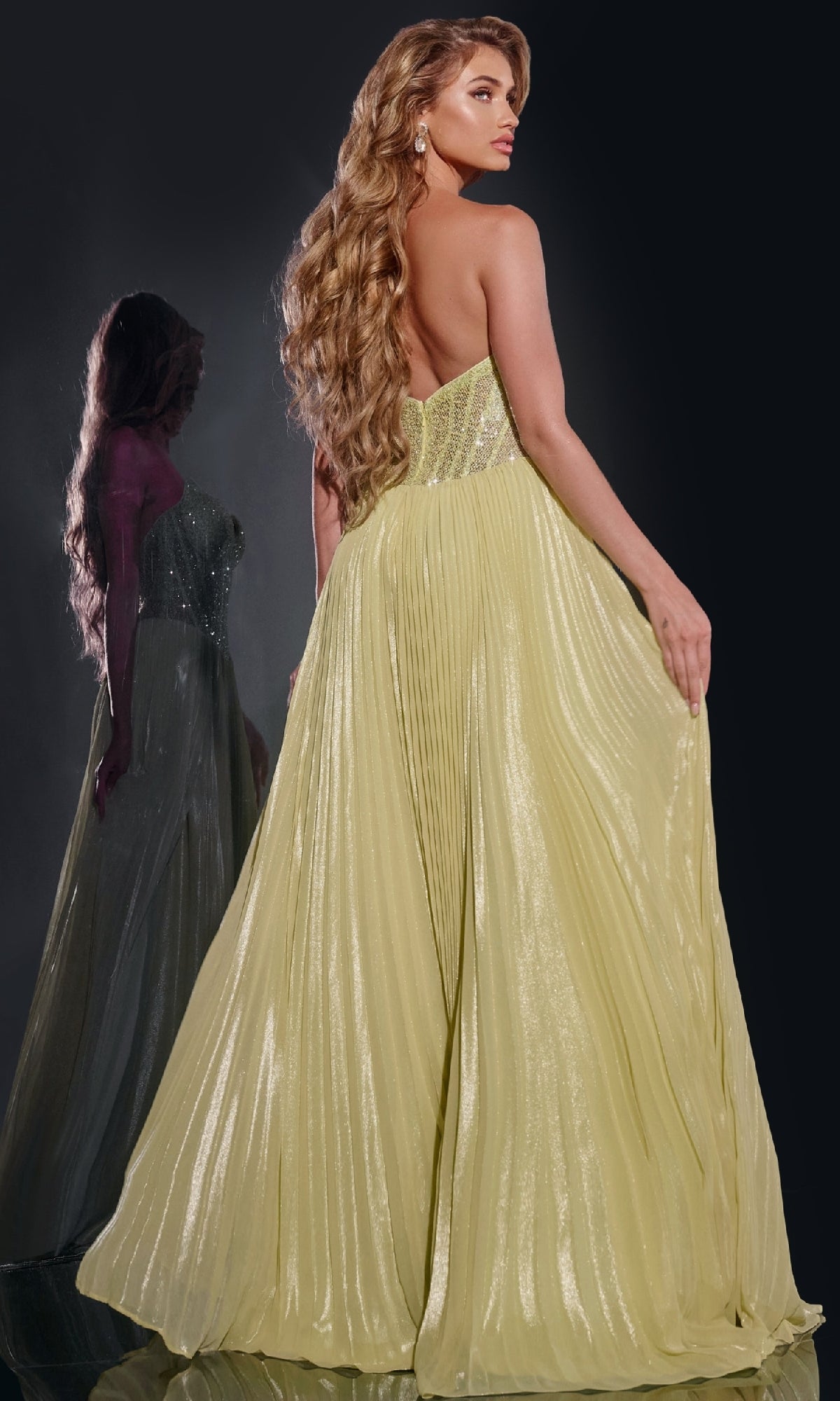 A woman wearing the Jovani 43115 long prom dress, featuring a shimmery strapless bodice and an A-line pleated skirt, stands with her back to the camera as her reflection appears in a mirror behind her.