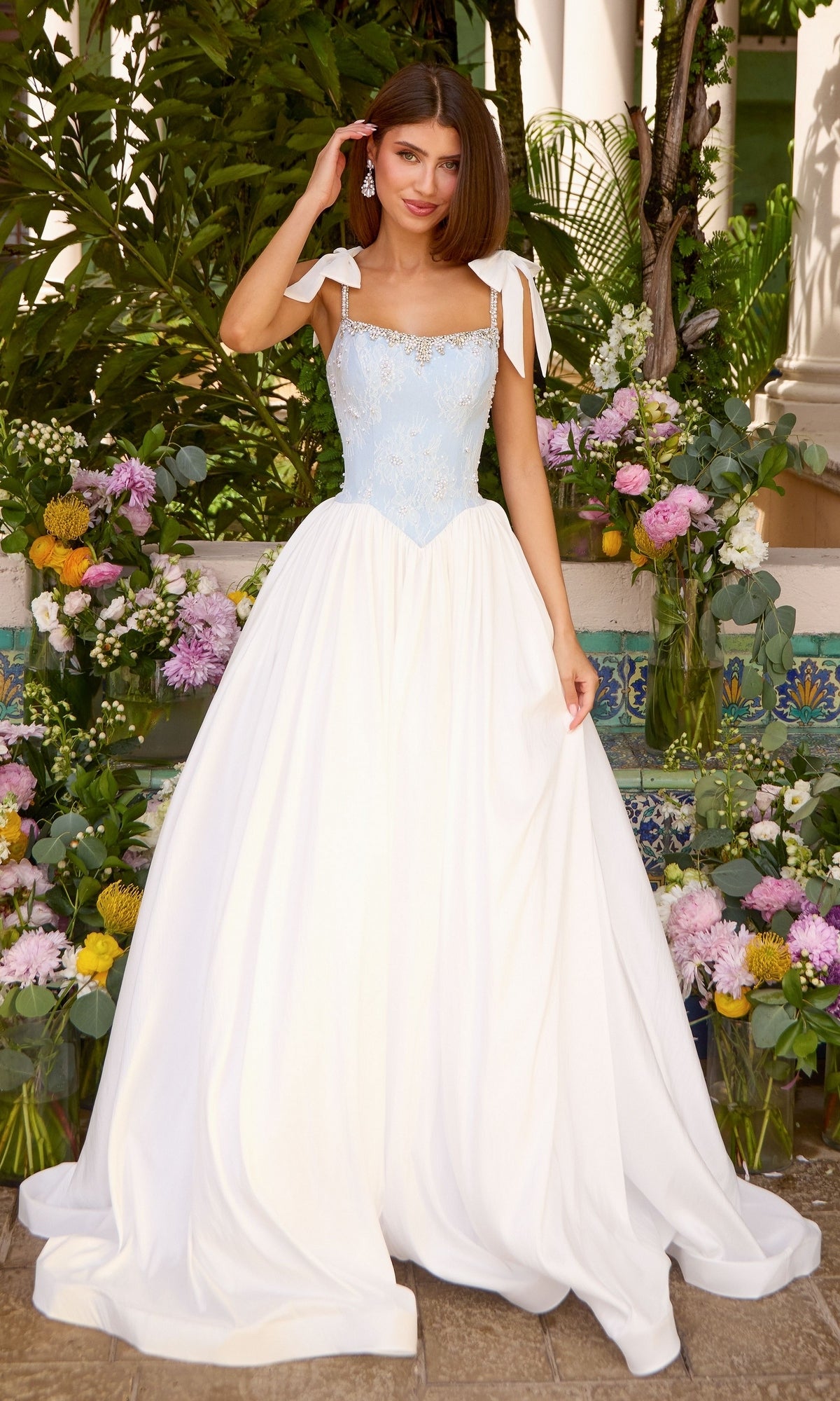 A woman wears an Ava Presley 43128 light blue prom dress with a beaded bodice, tie-shoulder straps, and a full white floor-length skirt while standing among arrangements of colorful flowers.