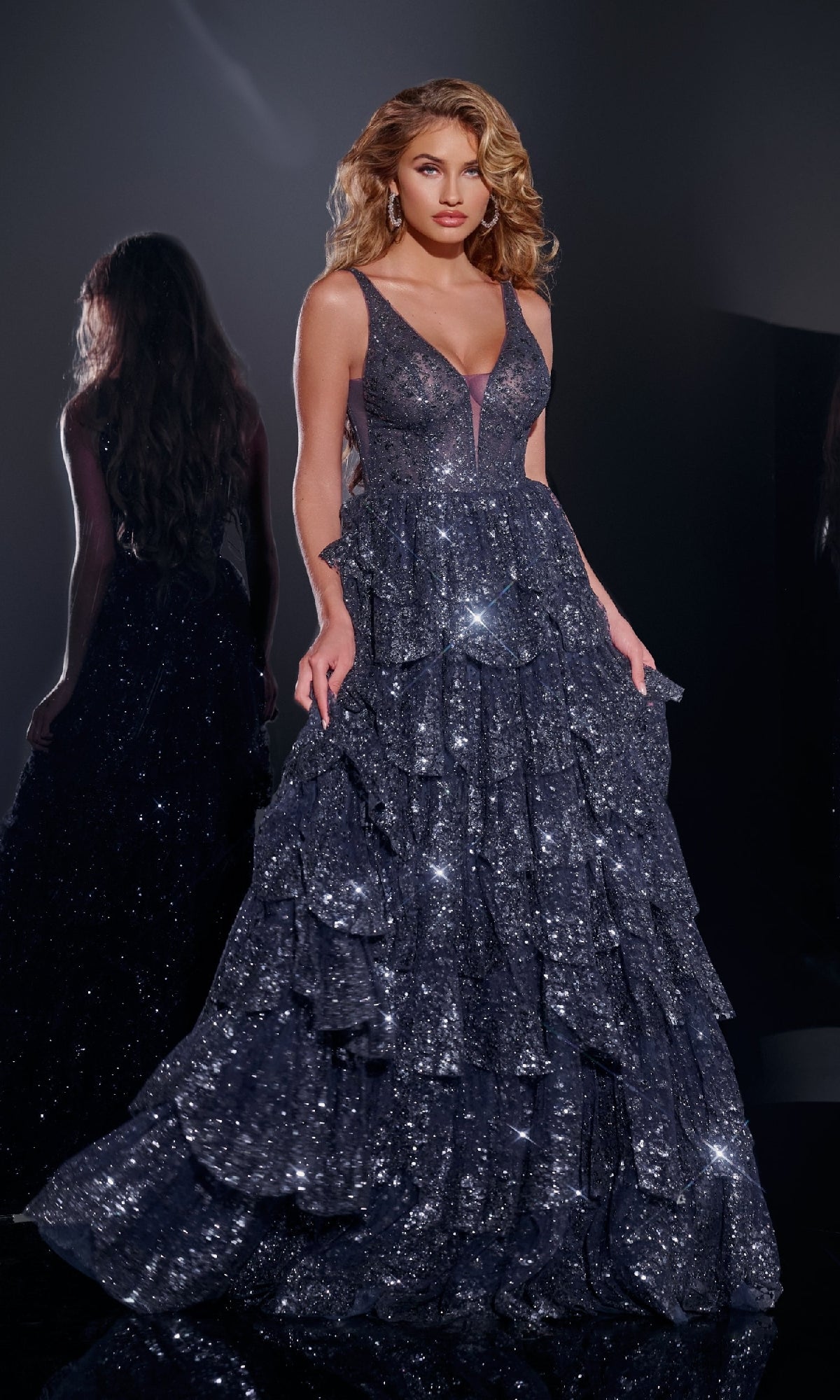 A woman stands in a dark room wearing the Jovani 43198 Ruffled Long Glitter Prom Dress in navy. The floor-length gown sparkles with layered ruffle details, her hair styled in loose waves, and her reflection visible behind her.