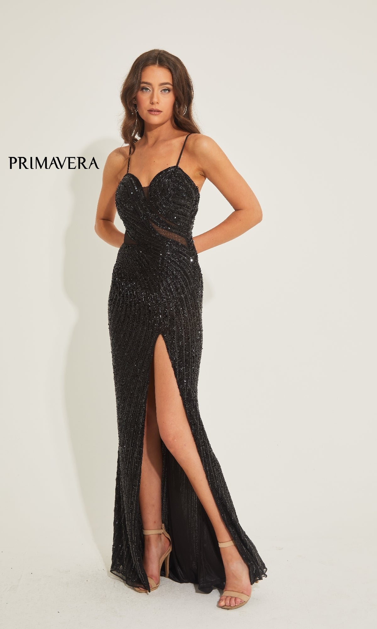 A woman models the Primavera 4327 long prom dress, a black sequined gown with spaghetti straps and a thigh-high slit, standing against a white background. The word