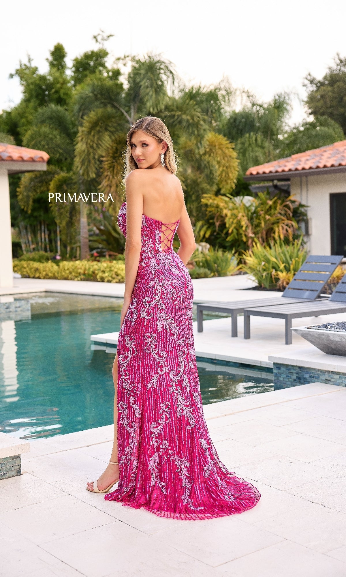 A woman in the Strapless Sequin-Print Prom Gown: Primavera 4328 stands by a pool facing away, with trees and modern buildings creating a striking backdrop for her shimmering formal dress.