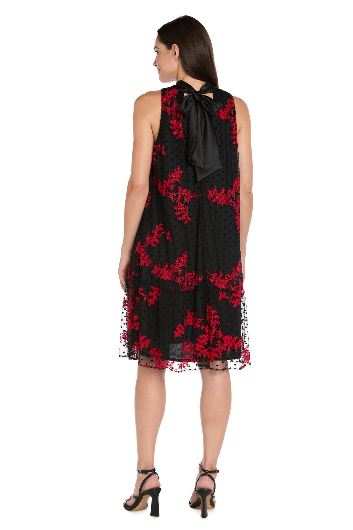 A woman faces away, wearing the Petite Short Black Party Dress R&M Richards 4328P—sleeveless, knee-length with red floral patterns, a black neck bow, and black heeled sandals.