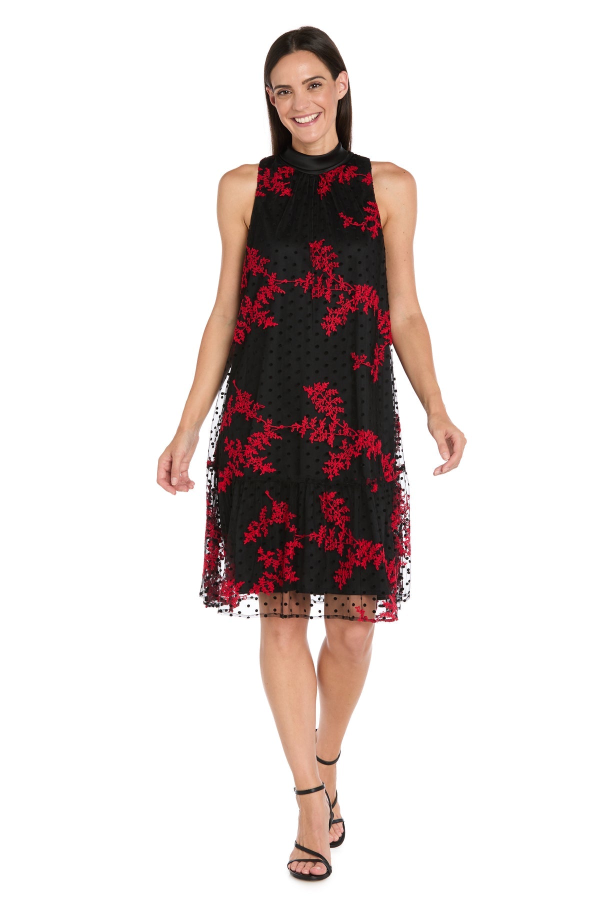 Woman in the R&M Richards 4328P Petite Short Black Party Dress (Black/Red), a sleeveless knee-length cocktail dress with red floral embroidery and sheer overlay, paired with black strappy sandals, standing and smiling.
