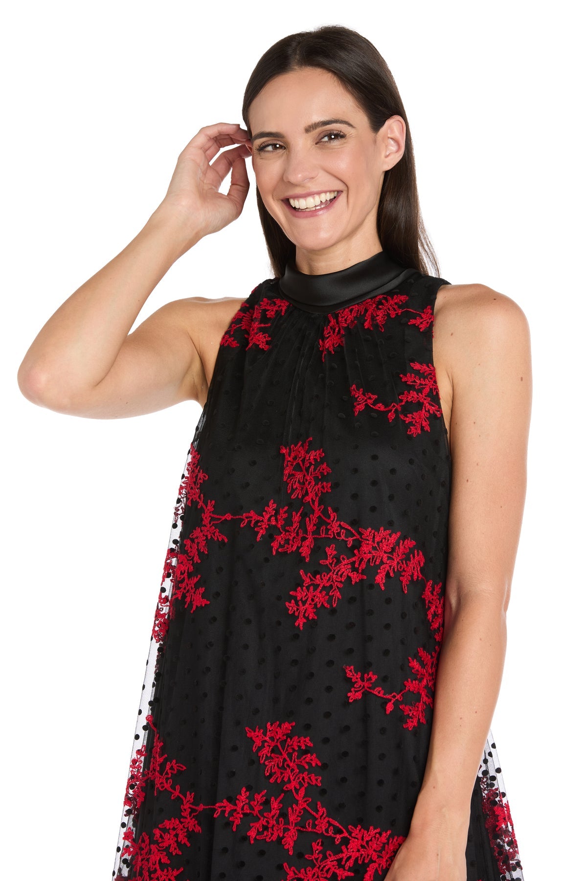 A woman smiles and touches her hair while wearing the R&M Richards 4328P Petite Short Black Party Dress with red floral patterns, posing against a white background.