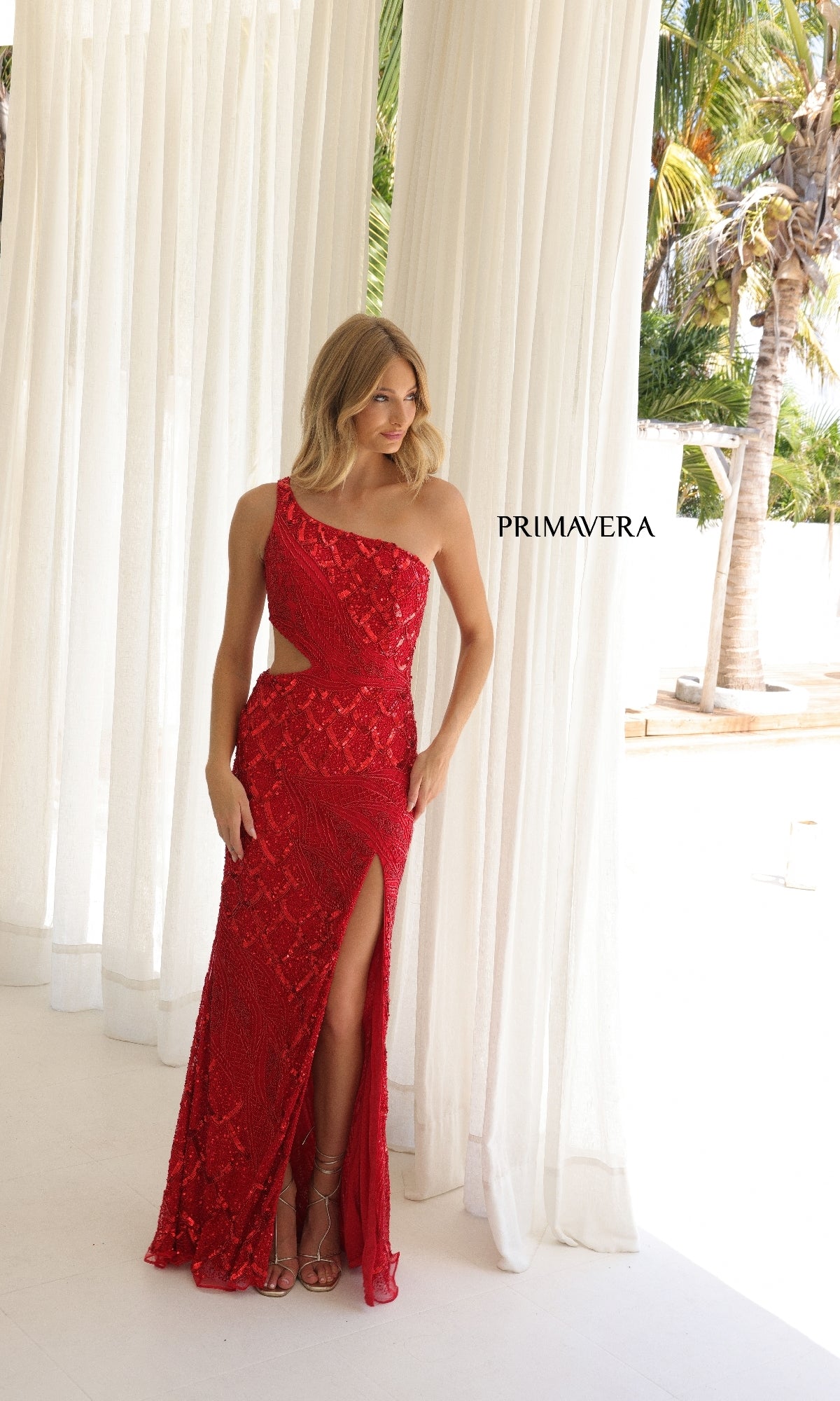 A woman stands indoors near white curtains and palm trees, wearing the Long Prom Dress: Primavera 4329—a red one-shoulder sequin gown with a thigh-high slit. “PRIMAVERA” is displayed beside her.