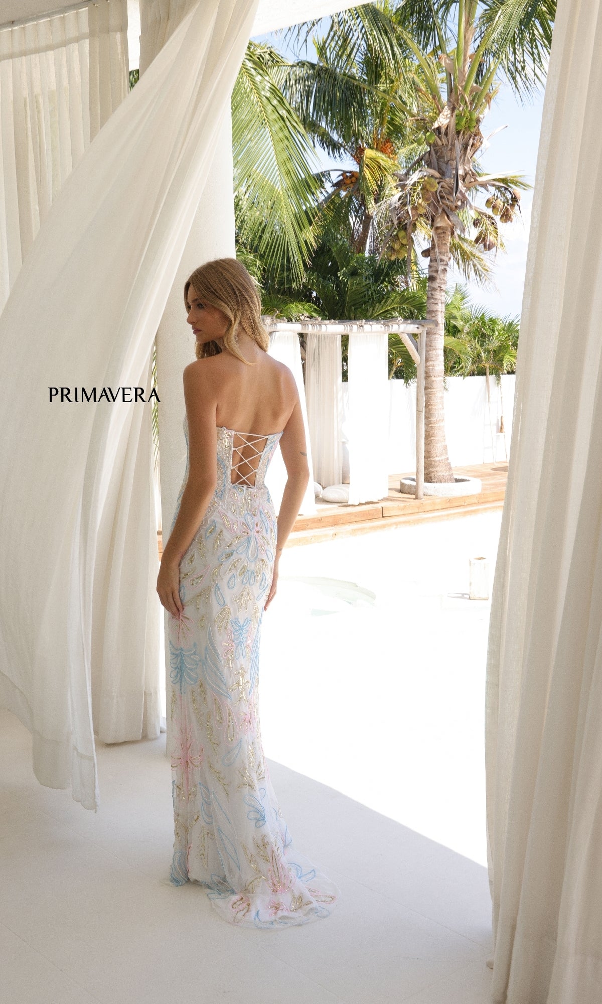A woman models the Strapless Beaded-Print Prom Dress: Primavera 4331 by white curtains, with palm trees and a pool behind her. The floor-length dress has a lace-up back, floral beading, and