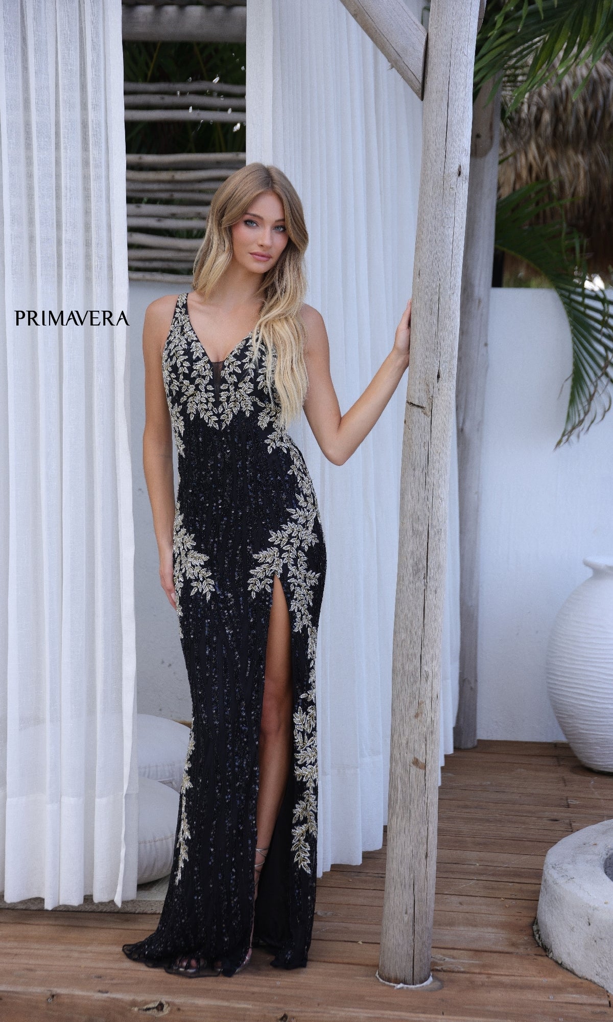 A woman stands on a wooden deck beside white curtains and a large vase, wearing the Strappy-Back Long Sequin Prom Gown: Primavera 4342, featuring gold embroidery, an open back, and a thigh-high slit.