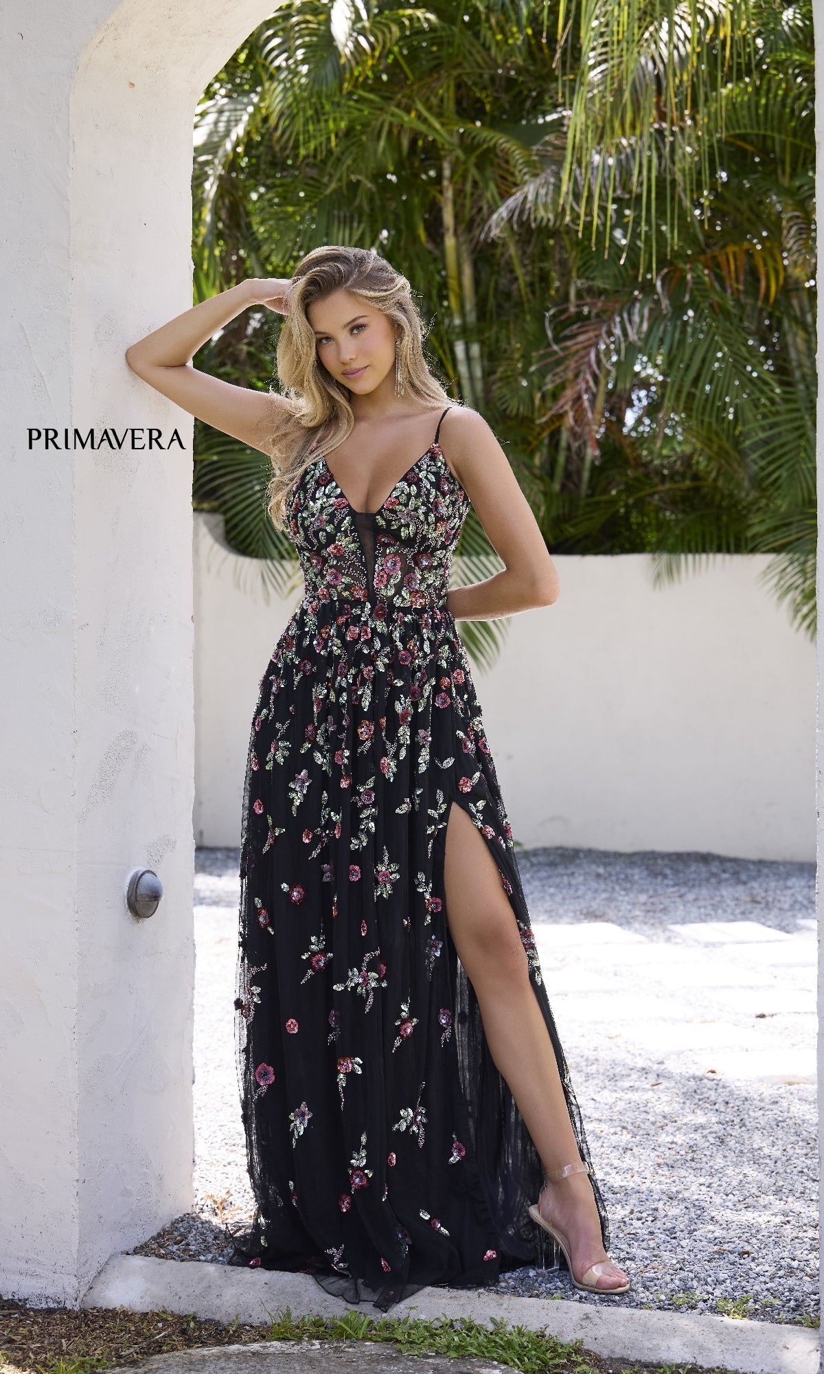 A woman stands outdoors against a white wall and lush green palms, wearing the Beaded-Floral Long Prom Dress: Primavera 4353—a black gown with floral embroidery and a high slit.