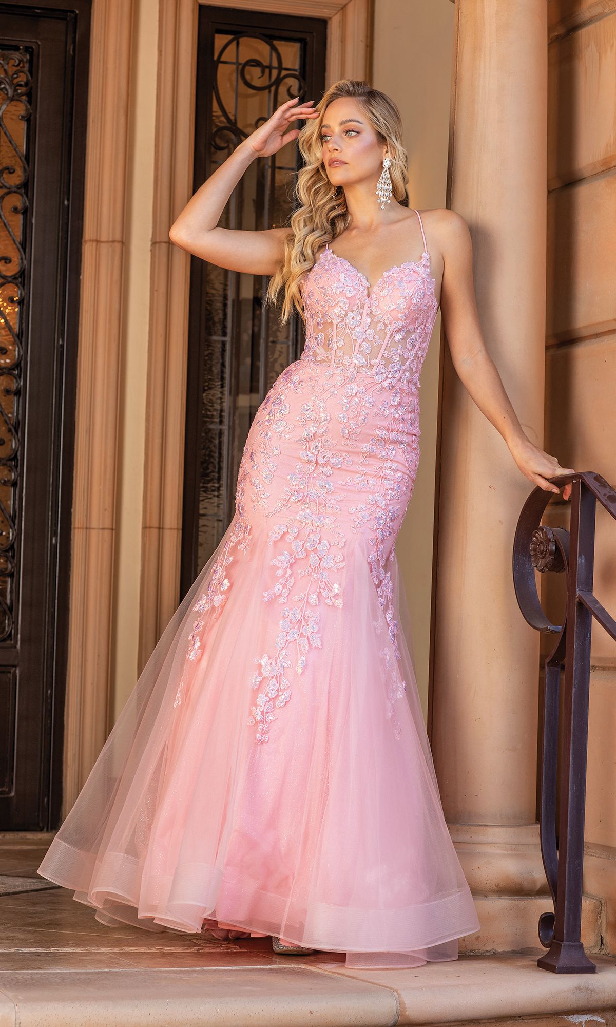 A woman stands on steps wearing the Backless Sheer-Corset Long Mermaid Prom Dress 4353, a fitted light pink gown with a sheer corset bodice and tulle skirt adorned with floral embellishments, holding the railing.