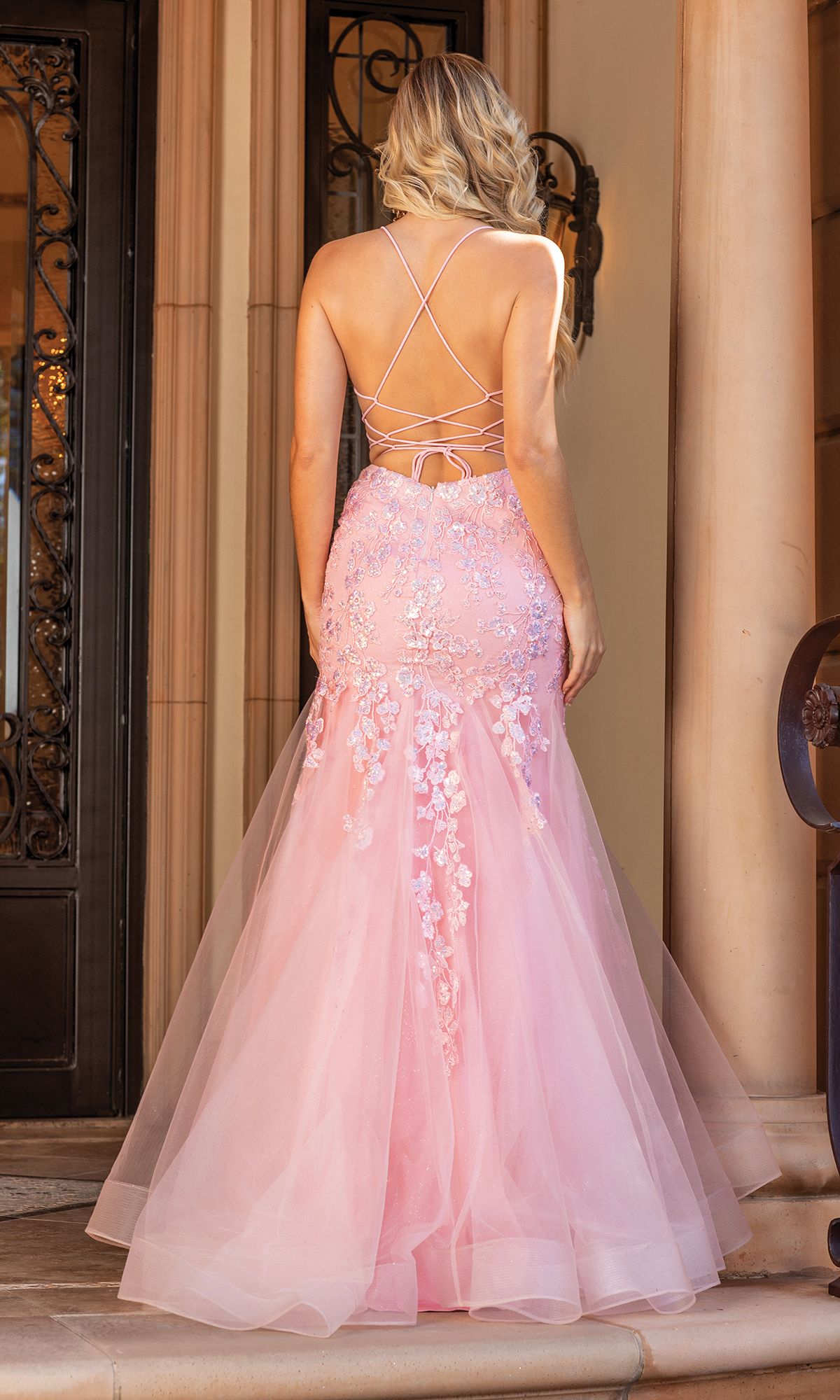 A woman stands on steps wearing the Backless Sheer-Corset Long Mermaid Prom Dress 4353, featuring light pink floral appliqué, embroidered lace details, and a lace-up open back.