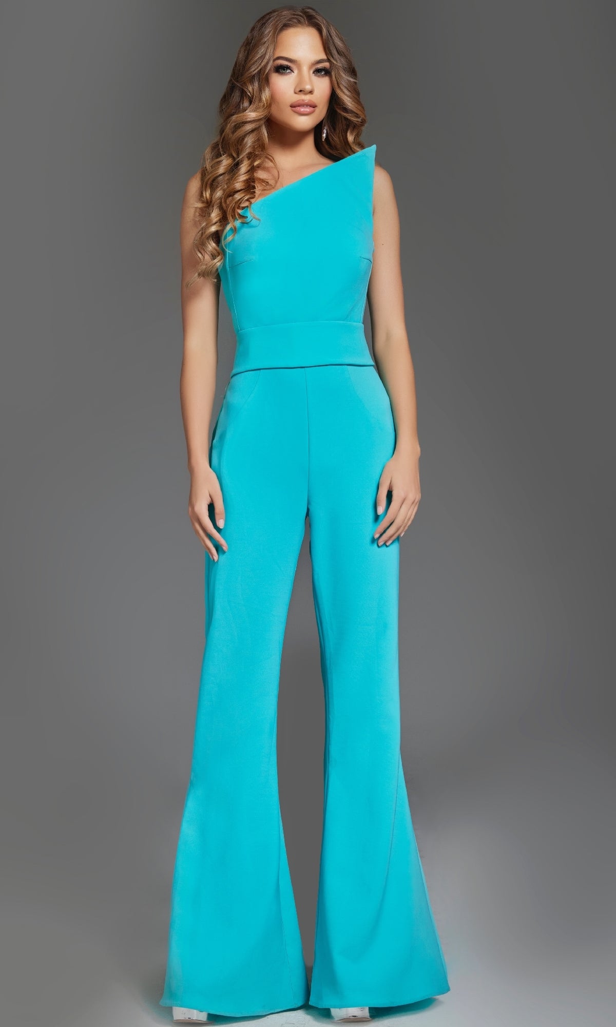 A woman models the Jovani 43567 Strapless Formal Jumpsuit in turquoise, featuring an asymmetrical strapless neckline and flared pant legs, set against a plain gray background.
