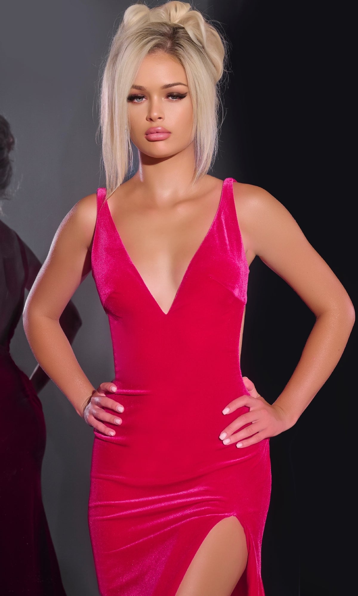 A blonde woman with a high bun models the Jovani 43578 long prom dress—a sleeveless pink velvet gown with a deep V-neckline and high slit—standing confidently with hands on hips against a dark background.