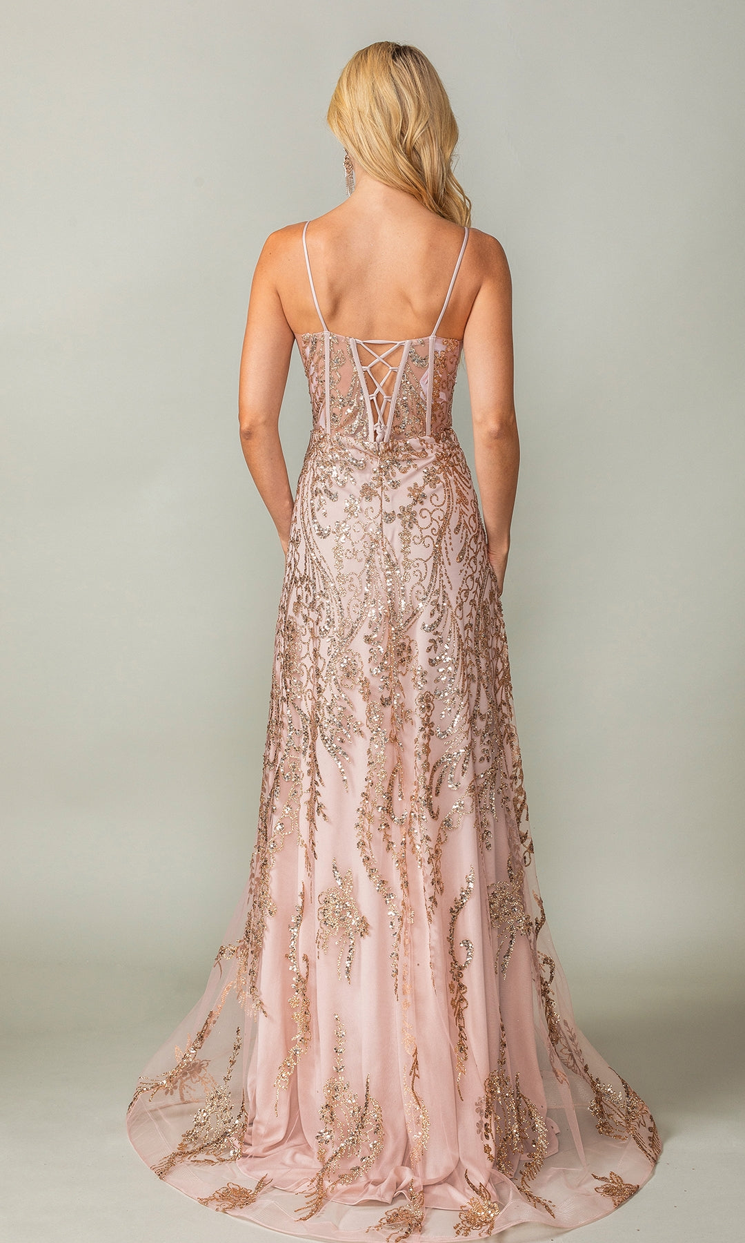 A woman stands facing away, wearing the Dancing Queen 4359 glitter-print long prom dress in pink, featuring thin straps, gold embroidery, and a lace-up back.