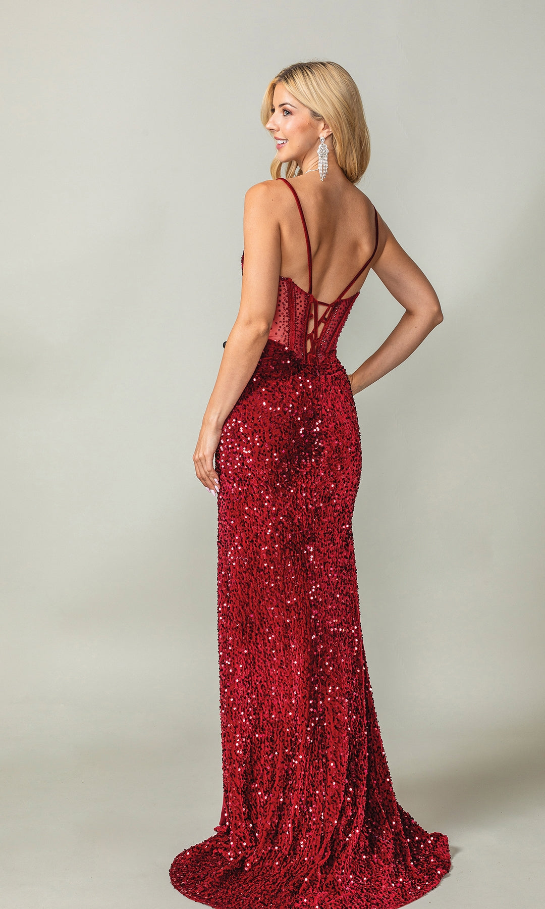 A woman poses and smiles in the Lace-Up Long Sequin Prom Dress: Dancing Queen 4360—a red, floor-length, sequin gown with thin straps and an open back—against a plain light gray background.