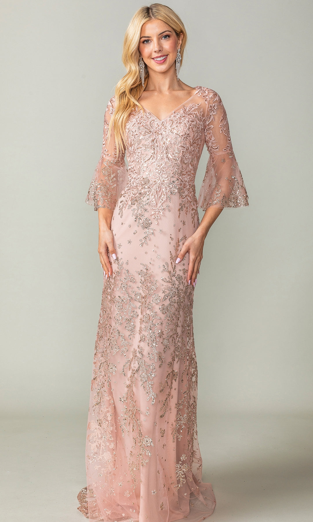 A woman wearing the Bell-Sleeve Long Prom Dress (Dancing Queen 4361)—a light pink lace gown with sheer bell sleeves and silver floral embroidery—stands against a plain background.