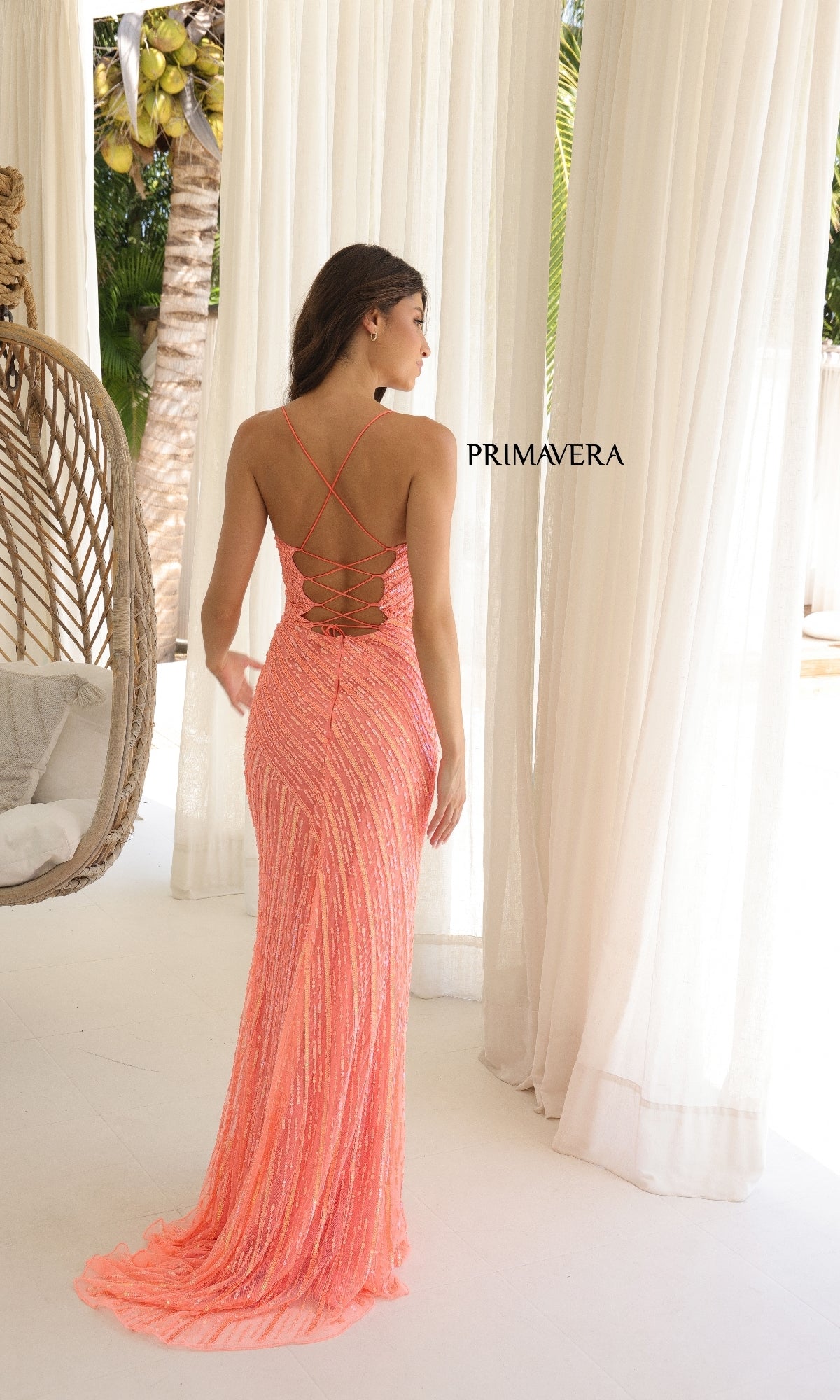 A woman stands indoors in the Strappy Long Beaded Prom Dress: Primavera 4363, a coral pink floor-length gown with crisscross straps and an open back. Light filters through sheer white curtains in the background.
