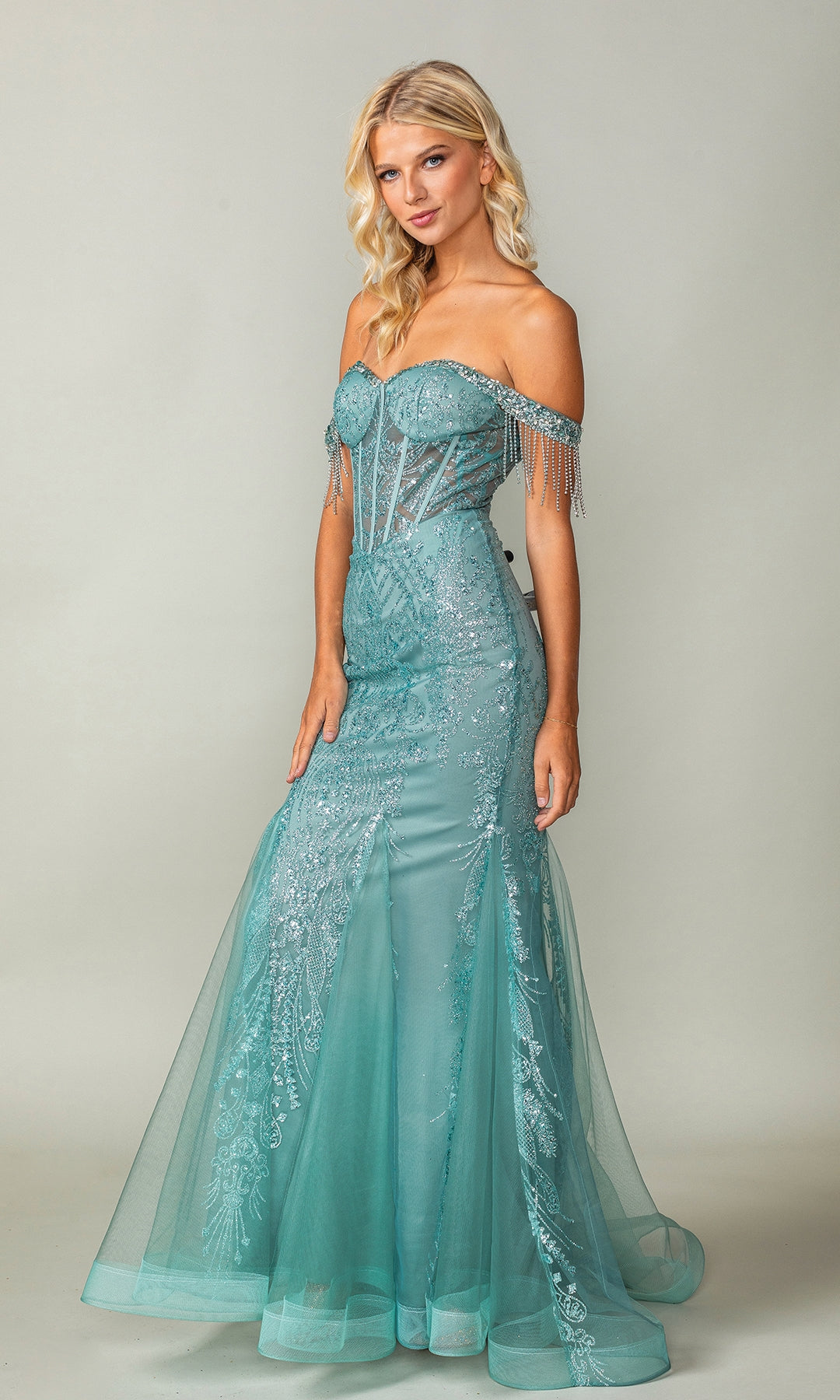 A woman models the Fringe Long Glitter Prom Dress: Dancing Queen 4365 in turquoise, showcasing an off-the-shoulder corset bodice with sheer details, sparkling beading, and a flared tulle skirt against a plain backdrop.