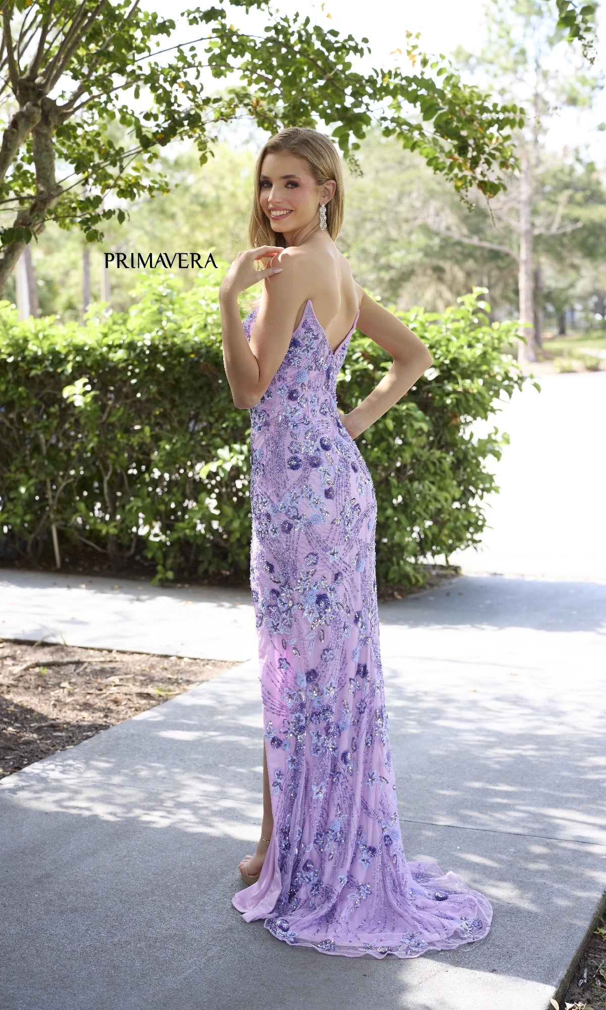 A woman poses and smiles outdoors in the Primavera 4374 long prom dress, featuring a strapless design with lilac floral embroidery and embellishments.
