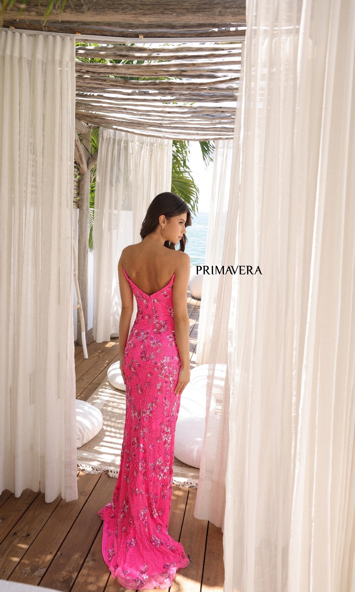 A woman wearing the Primavera 4374, a bright pink floral long prom dress, stands with her back to the camera on a wooden deck framed by sheer white curtains.