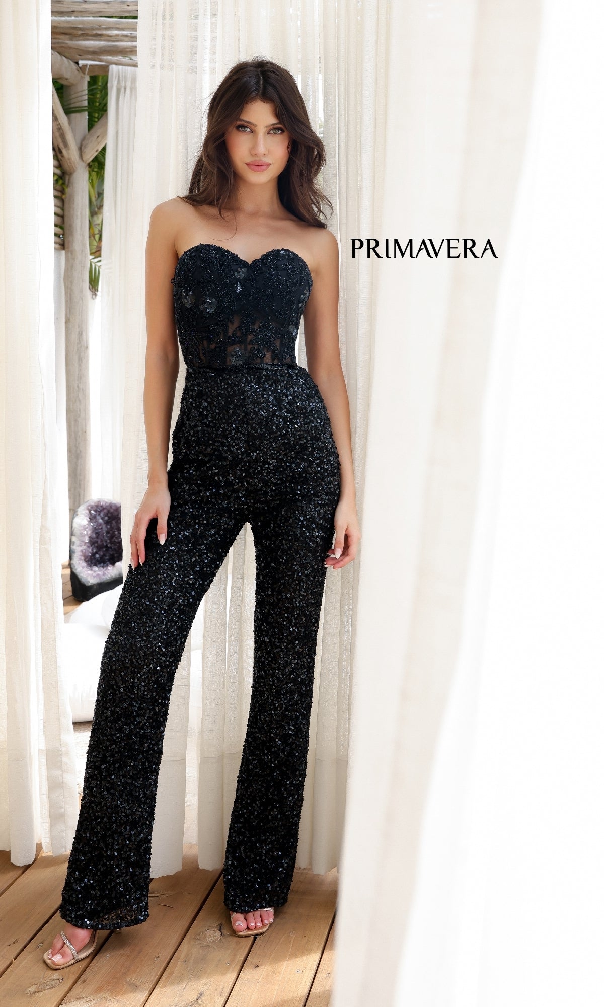 A woman stands indoors in the Long Prom Jumpsuit: Primavera 4383 with black sequins and heels, framed by sheer white curtains and wooden flooring.