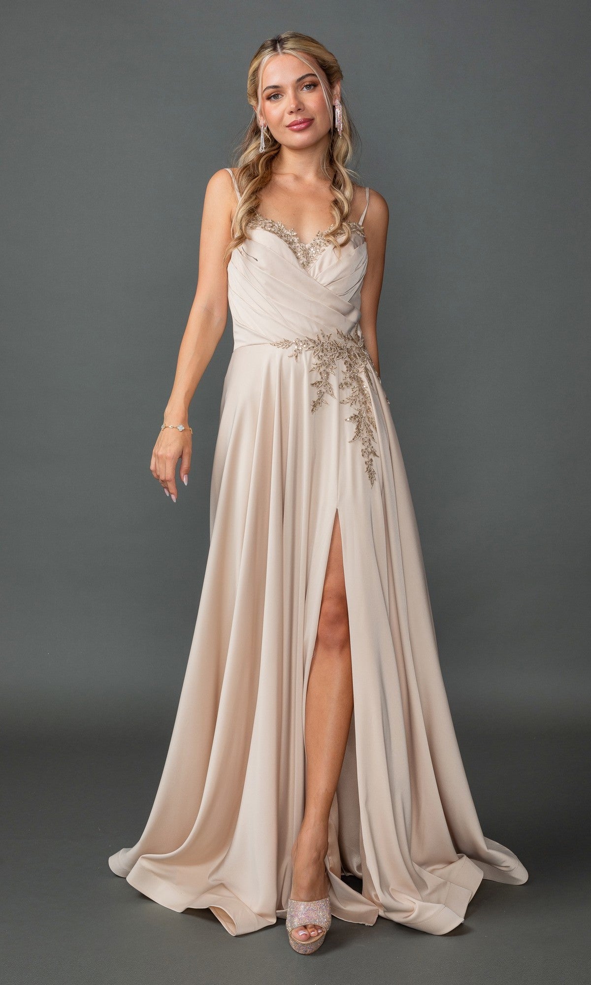 A woman stands against a plain gray background wearing the Lace-Up-Back Long A-Line Prom Dress 4391 in champagne, featuring embroidery, lace detailing, a thigh-high slit, and spaghetti straps.