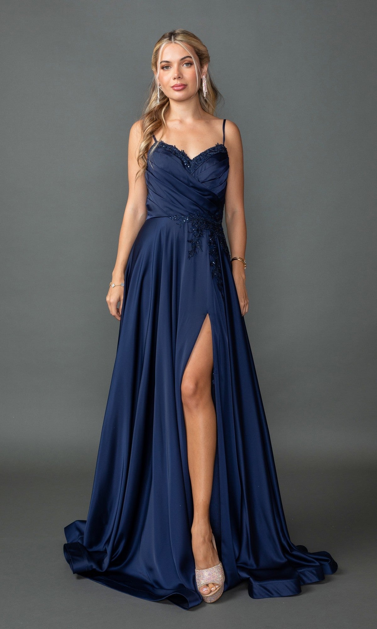 A woman models the Lace-Up-Back Long A-Line Prom Dress 4391 in navy, featuring a thigh-high slit, spaghetti straps, and floral embellishments, while standing against a plain gray background.