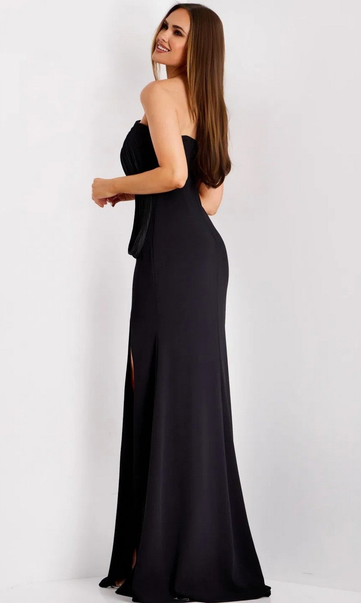 A woman wearing the Jovani 43970 Long Prom Dress—a chic, strapless black gown with a side slit—stands against a white background, glancing over her shoulder with a smile.