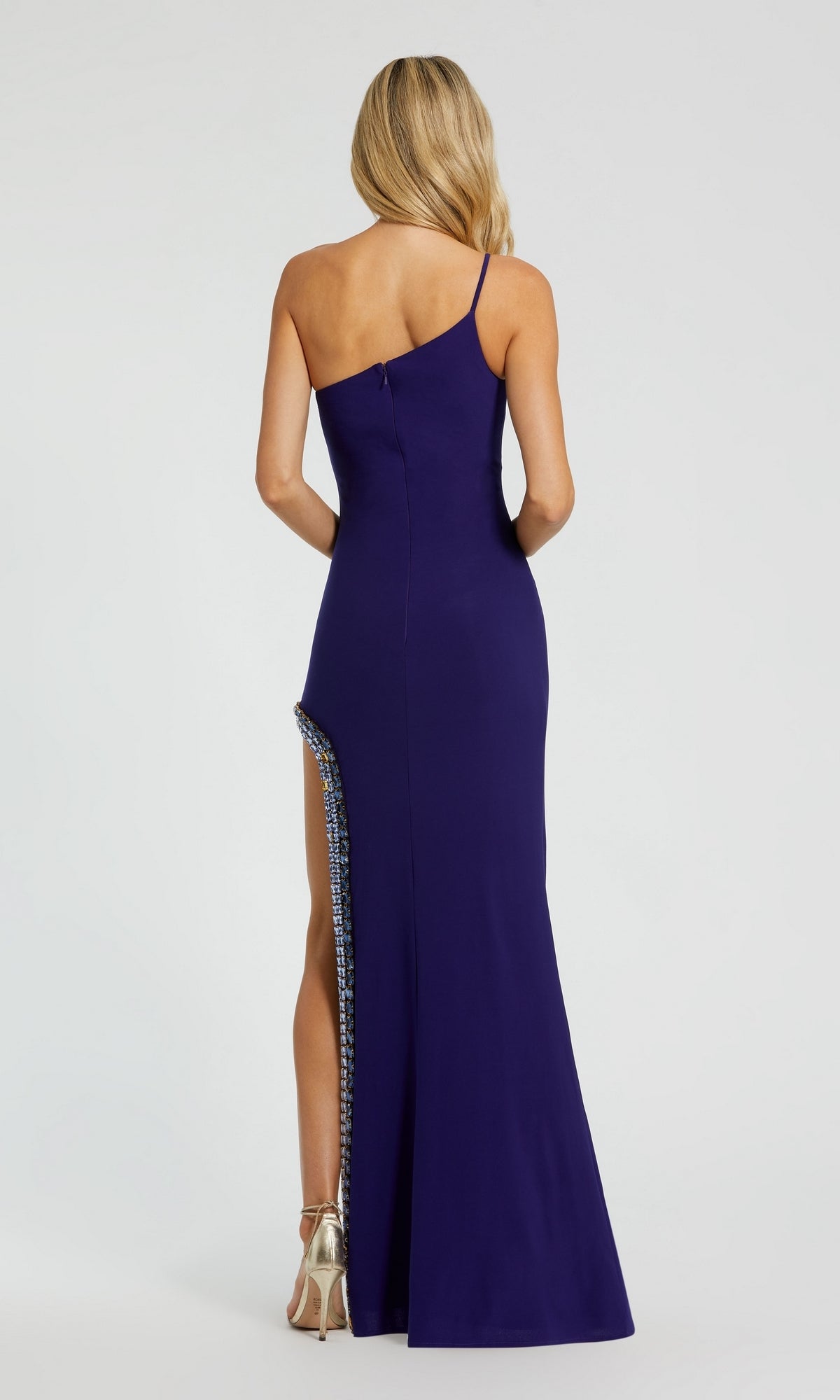 A woman stands facing away, wearing the Mac Duggal 44030 Bead-Trim Long Indigo Prom Dress with a one-shoulder design and high side slit, paired with metallic heels.