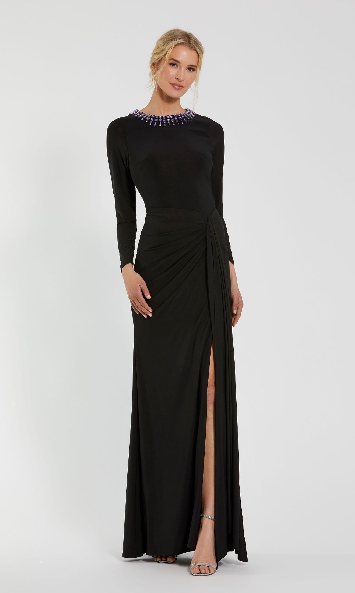 A blonde woman wears the Mac Duggal 44069 Bead-Neckline Long Formal Dress—a modest, long-sleeve gown with a high slit, gathered side, and beaded neckline. She pairs it with strappy heels.