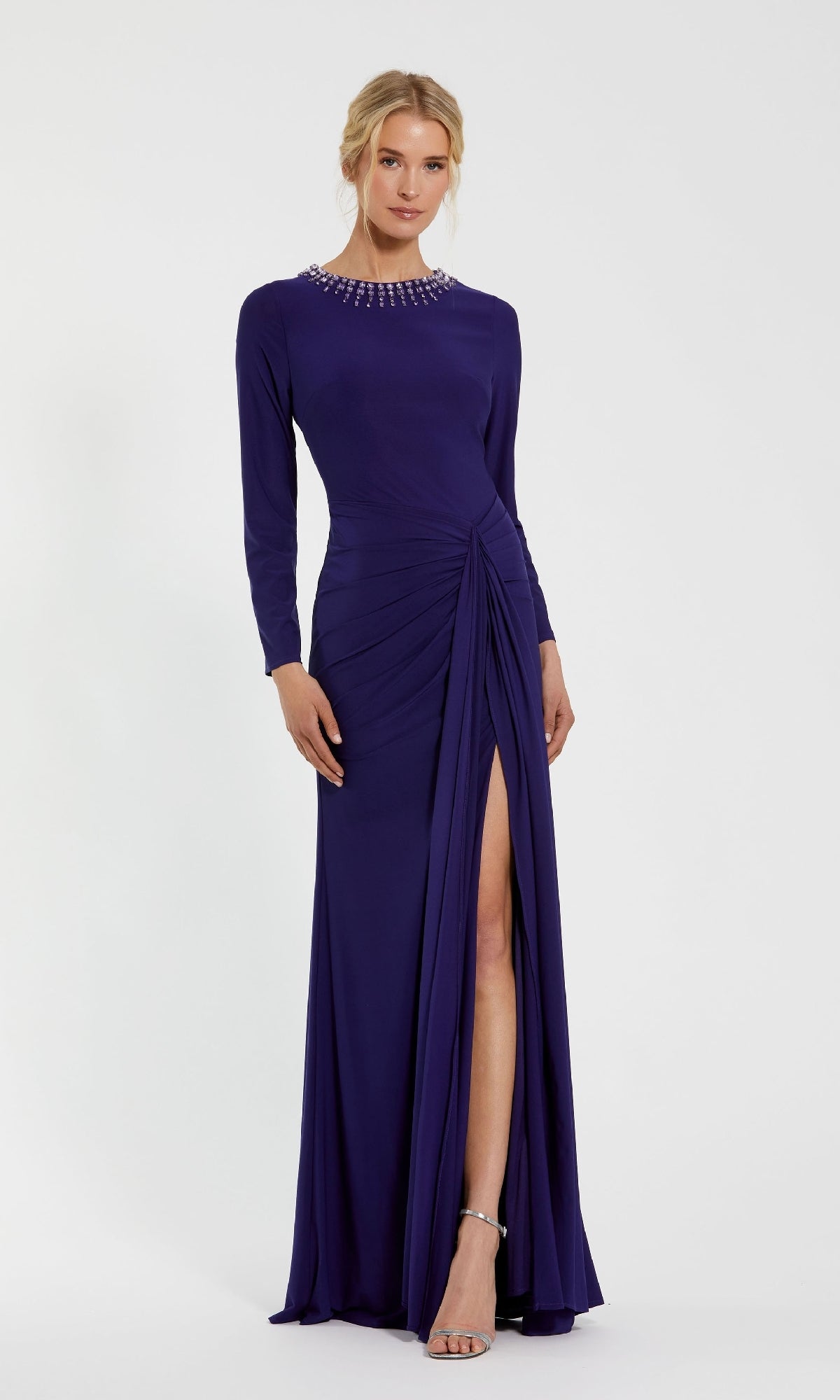 A woman in the Mac Duggal 44069 Bead-Neckline Long Formal Dress—a floor-length purple gown with long sleeves, a thigh-high slit, and pearl-embellished neckline—wears silver open-toe heels and stands against a plain white background.