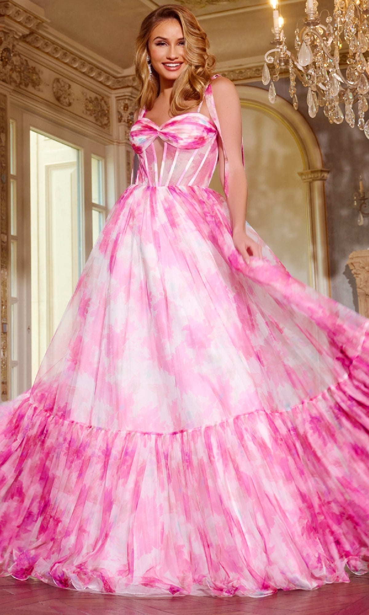 Woman wearing the Jovani 44181 Watercolor-Print Pink Prom Gown, featuring a ruffled hem, poses indoors in an elegant chandelier-lit room.