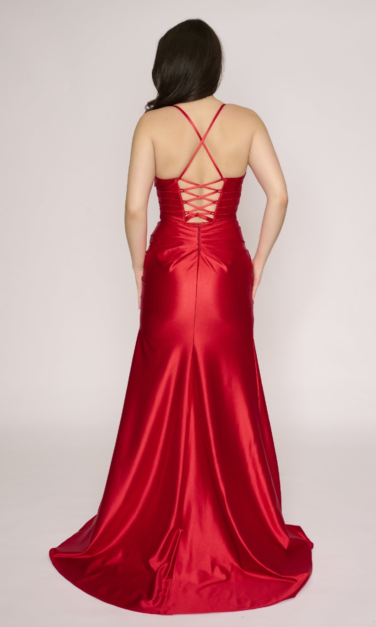 Woman wearing the Nina Canacci 4426 long formal dress in red satin, with a fitted bodice, v-neckline, criss-cross straps, lace-up back, and chic thigh-high slit, facing away from the camera.