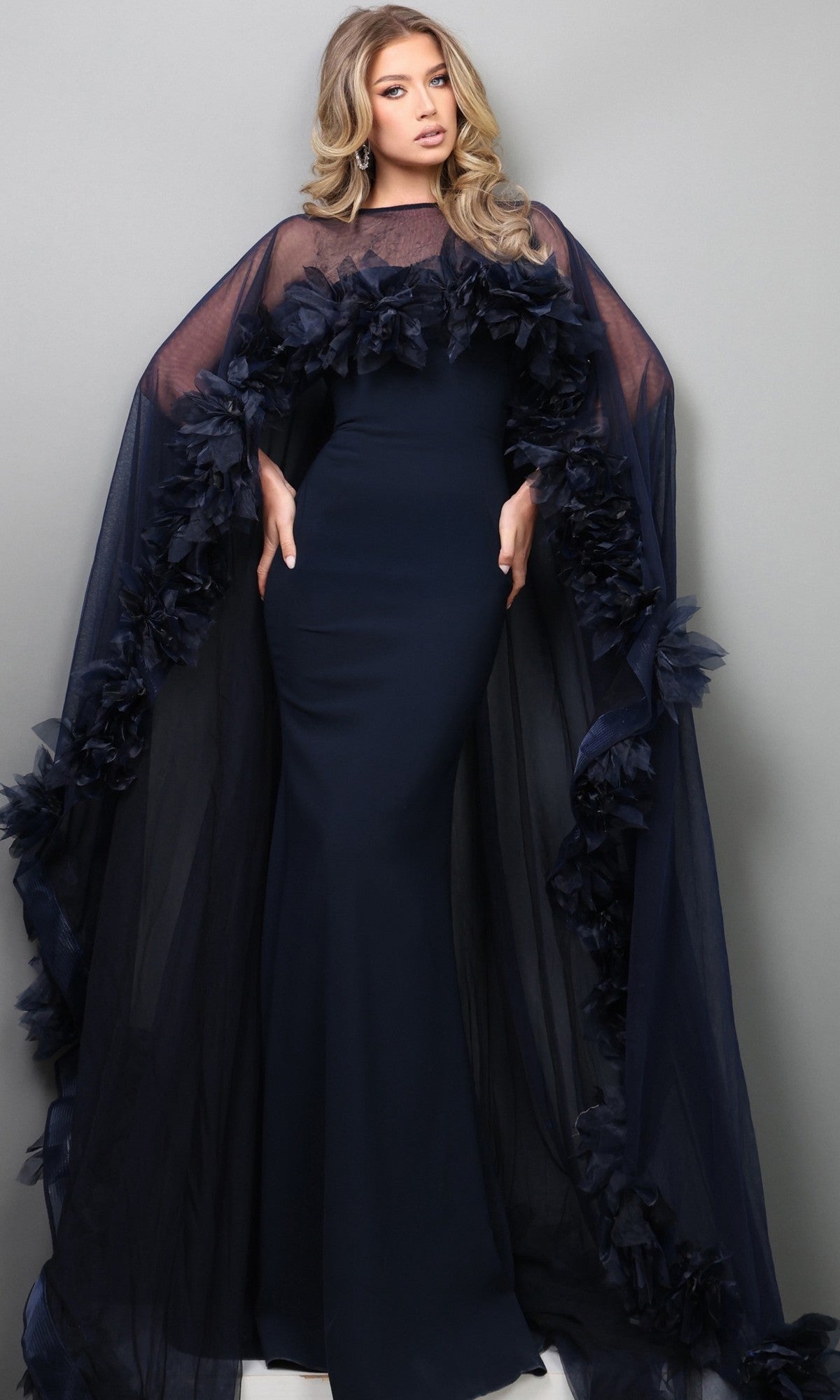 A woman stands against a gray background, hands on hips and looking forward, wearing the Long Formal Dress: Jovani 44321—a fitted navy blue designer gown featuring a sheer, ruffled cape.