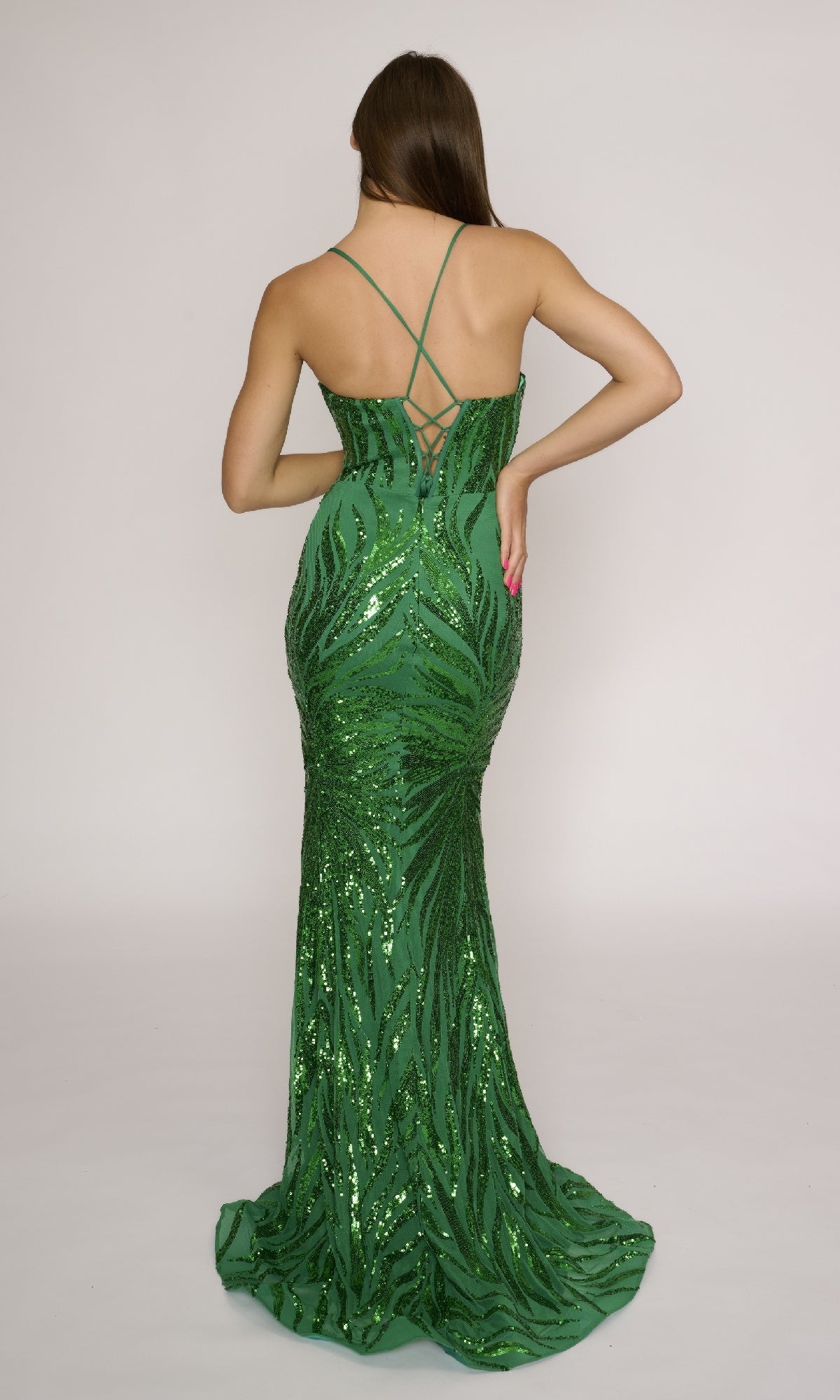 A woman faces away in the Nina Canacci 4433, a fitted green sequin prom dress with thin crisscross straps, a lace-up open back, and an elegant long flowing hem.