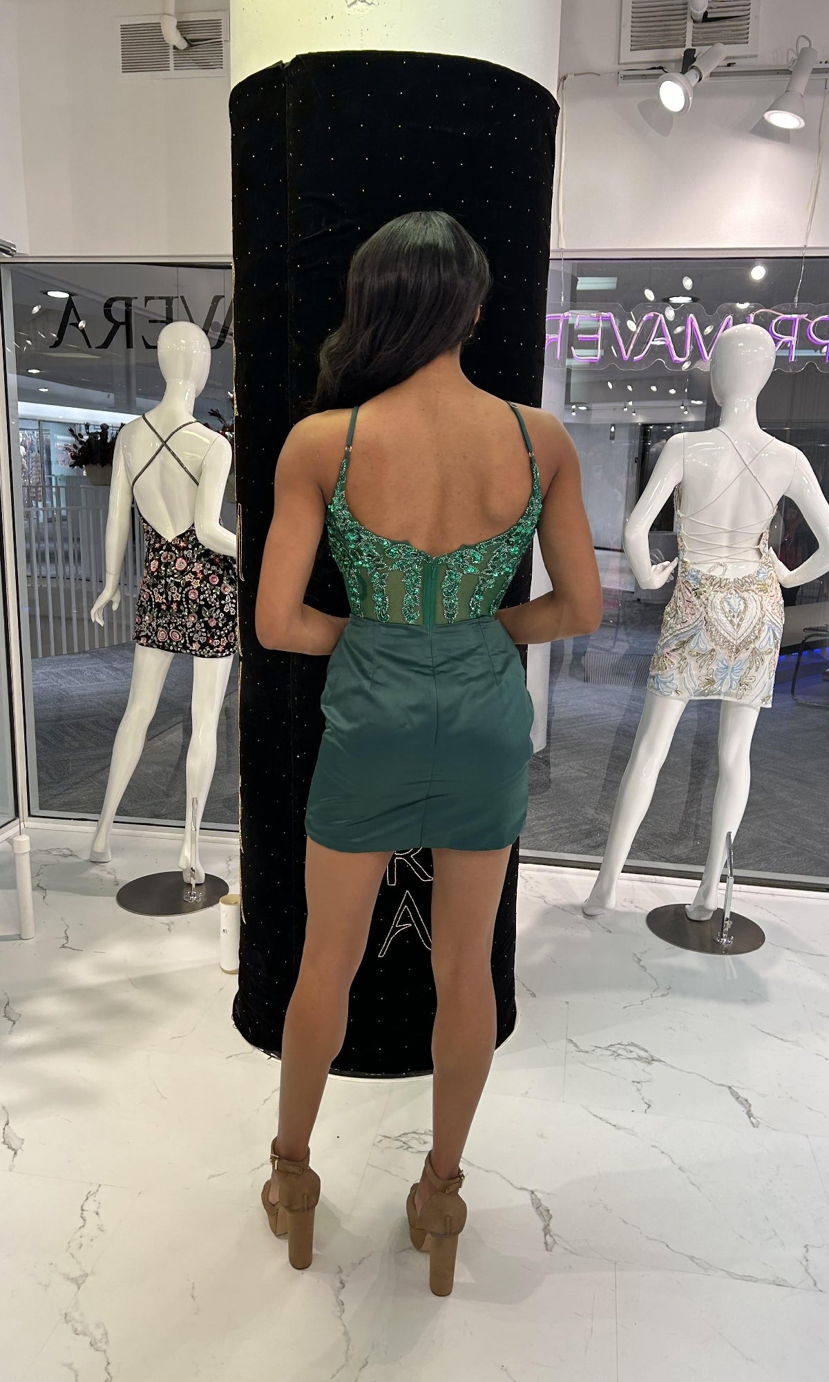 A woman in a green satin wrap skirt with a lace top and tan platform heels stands facing away; in front of her, two mannequins display sleeveless Short Homecoming Dresses: Primavera 4437.