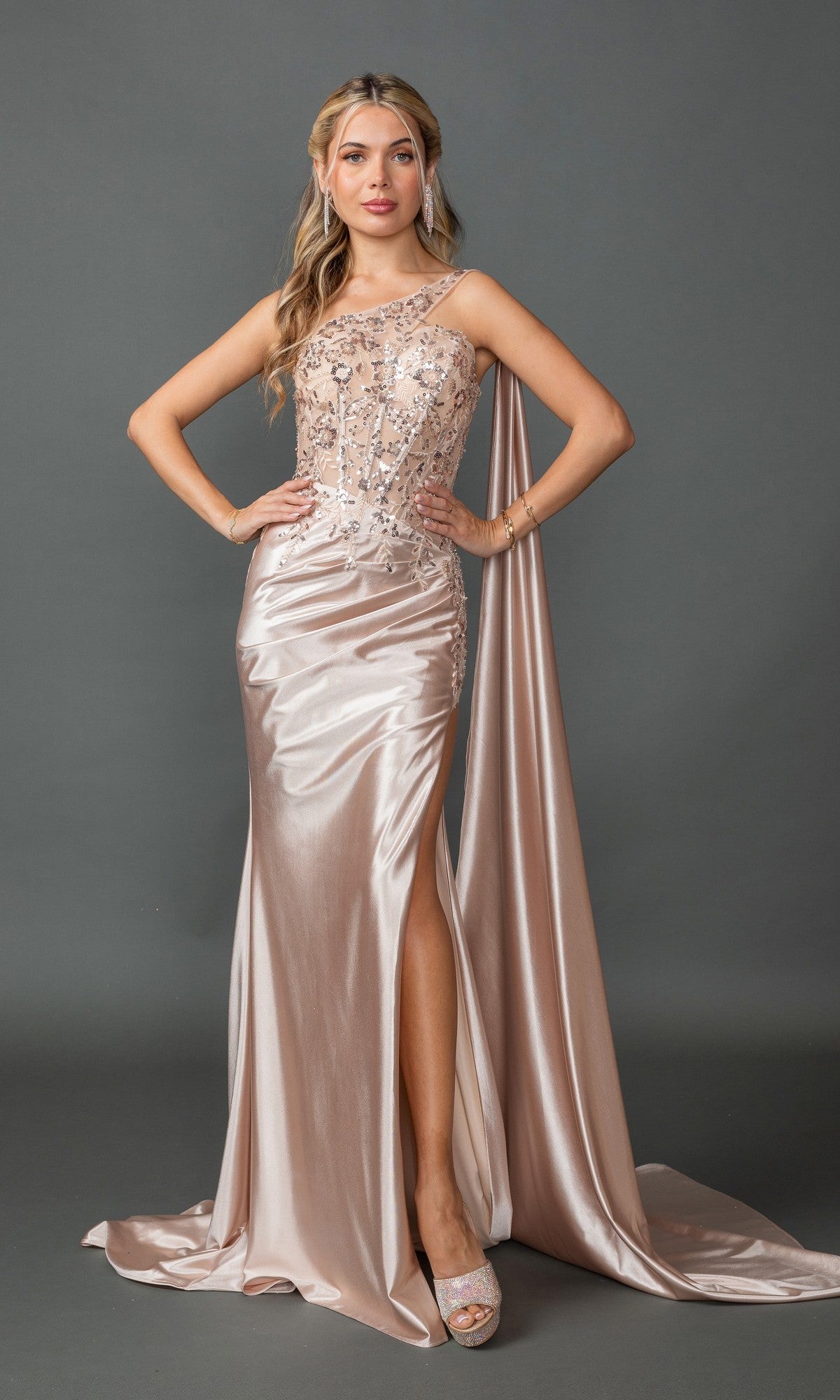 A woman poses against a plain gray background in the Dancing Queen 4441 one-shoulder long prom dress in champagne, featuring beaded lace, a sheer embellished bodice, thigh-high slit, and flowing cape.