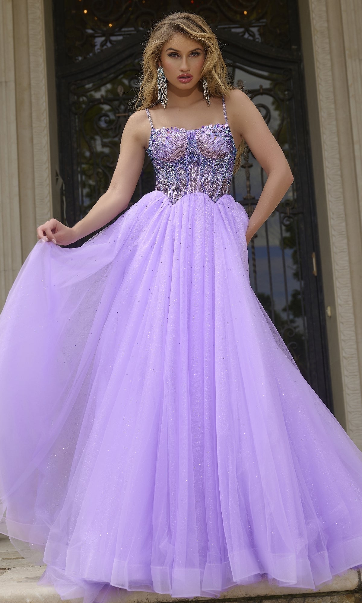 A woman stands before ornate doors in the Jovani 44511 Beaded-Corset Long Prom Ball Gown—a lavender creation with a beaded bodice and tulle skirt—holding out one side of her dress.