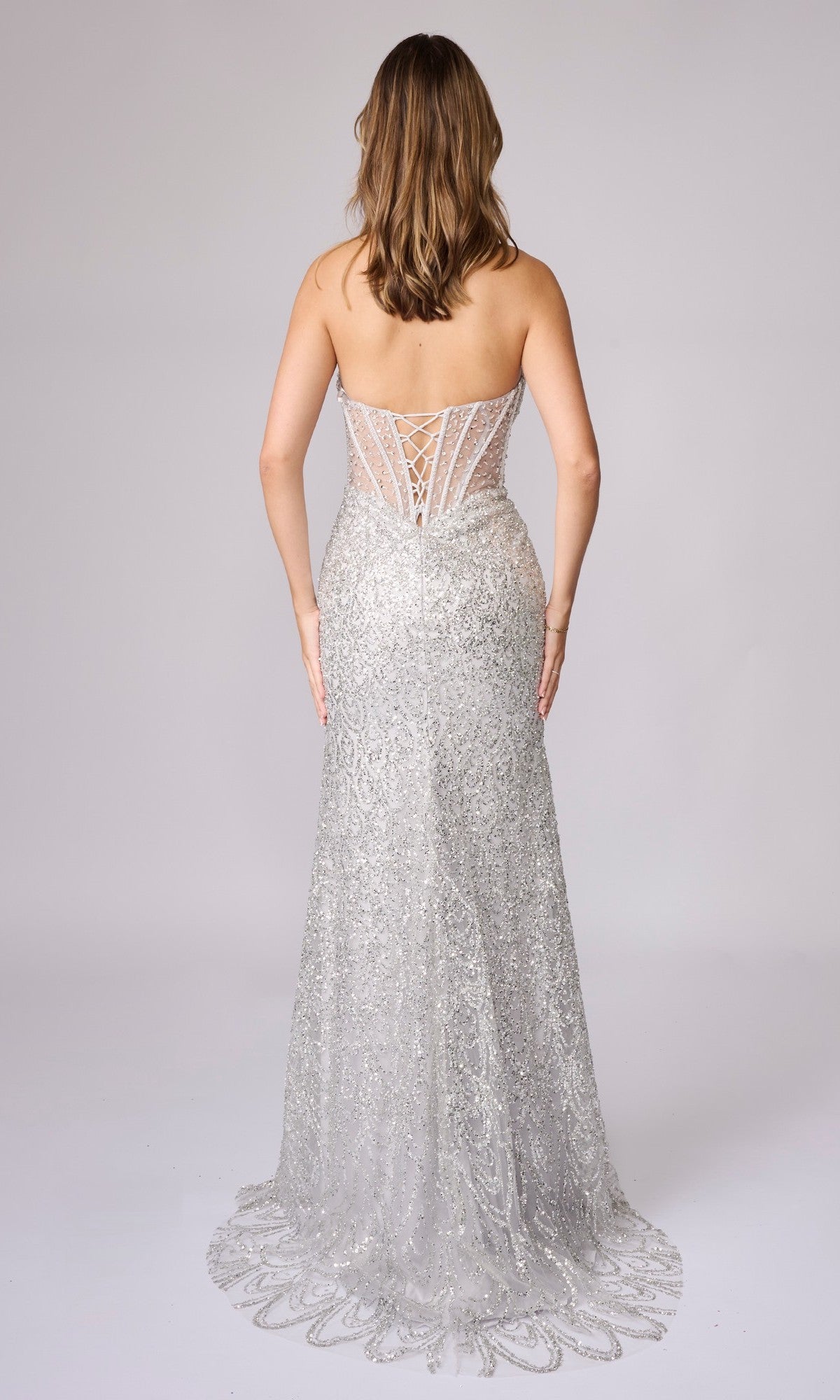 A woman faces away, wearing the Long Sequin Prom Dress: Nina Canacci 4458—a silver, strapless gown with a sheer corset bodice, embellished back panel, and sparkling sequined floor-length train.
