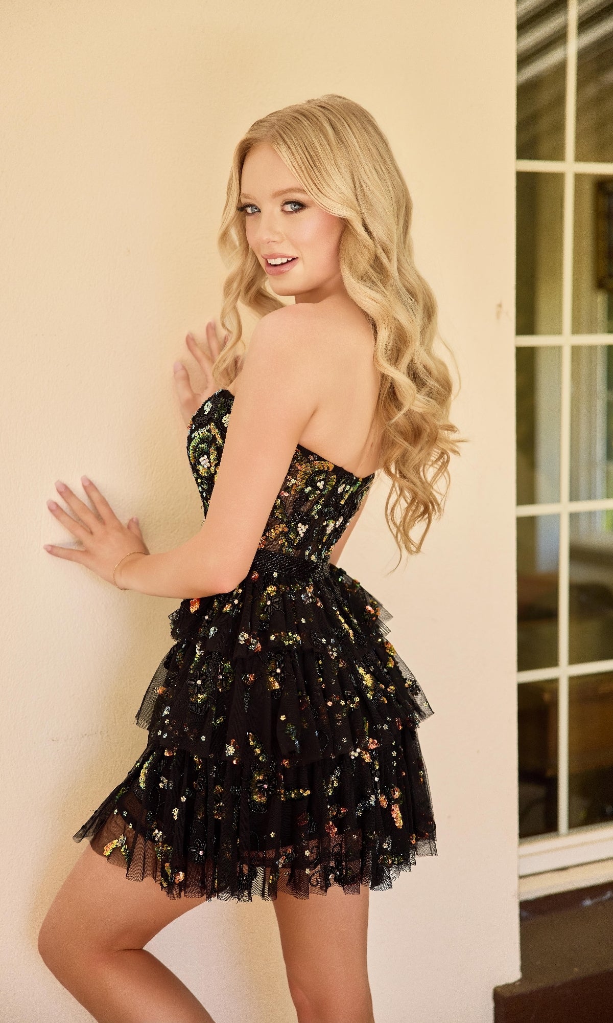A young woman with long blonde hair poses indoors against a light-colored wall, wearing the Primavera 4463 short homecoming dress—a black, strapless fit-and-flare style with a floral pattern.