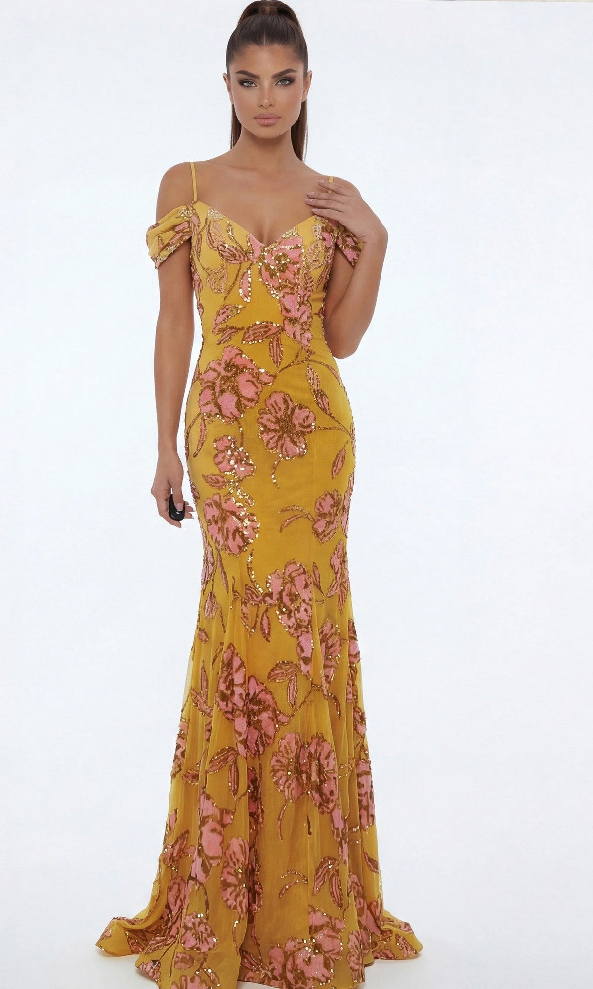 A woman models the Jovani 44652 long formal dress, featuring a yellow base with pink floral patterns, as she stands against a plain white background facing forward, one hand at her side and the other touching her hair.