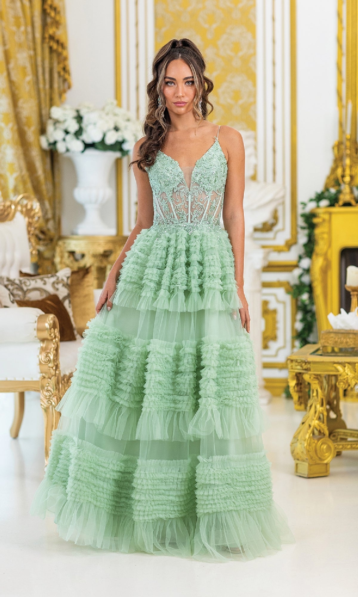 A woman stands indoors in the Sage Green Ruffled Prom Ball Gown (Dancing Queen 4476), featuring a lace bodice and tiered ruffles; ornate gold and white decor adorns the background.