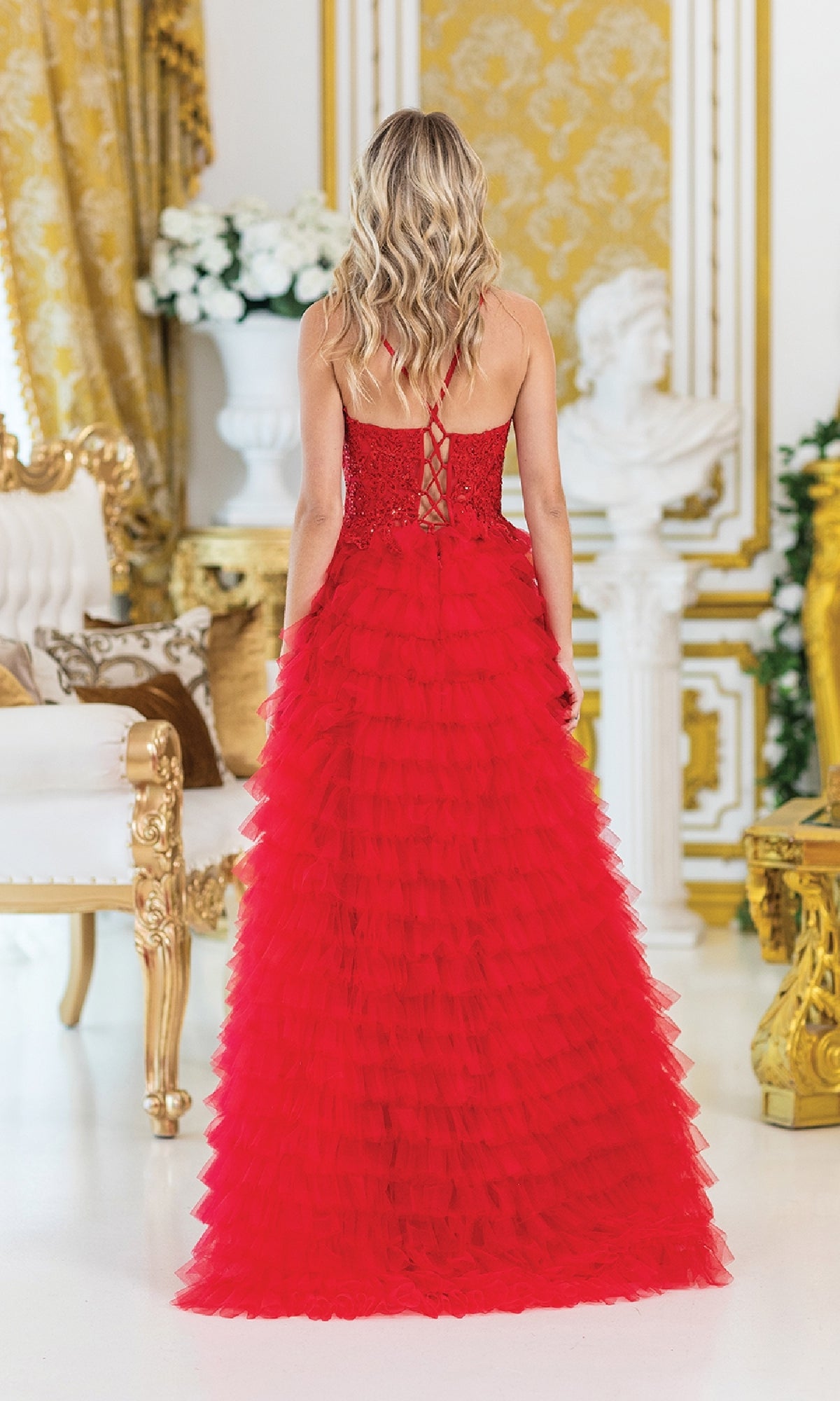 A woman with long wavy blonde hair stands in an ornate room, wearing the red, floor-length Dancing Queen 4491 Long Ruffled Prom Ball Gown with a lace-up back, giving her an elegant look as she faces away from the camera.