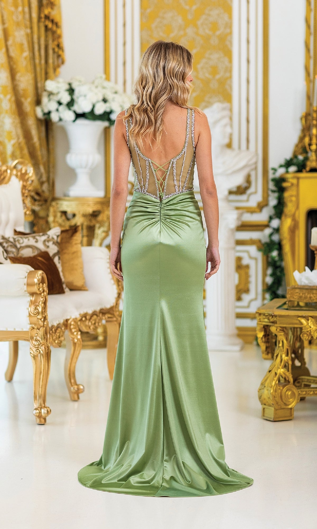 A woman stands in an ornate room wearing the Sheer-Corset Long Prom Dress: Dancing Queen 4505—a light green satin gown with a beaded sheer back, corset-style bodice, and elegant ruching.