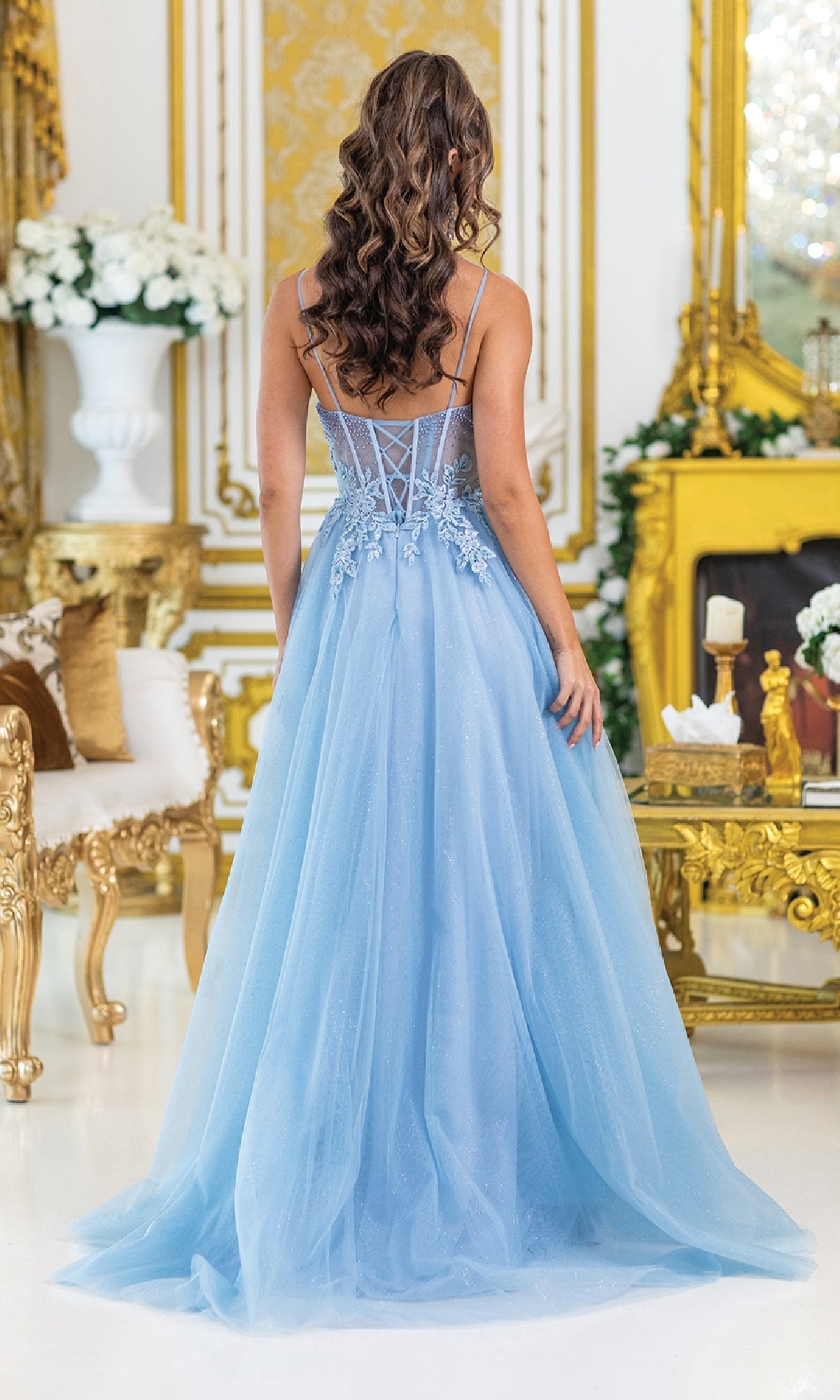 A woman with long, wavy hair wears the Corset Prom Ball Gown with Slit: Dancing Queen 4529—an embroidered light blue gown with a sheer back and floral details—while standing in an ornate room with gold accents and white flowers.