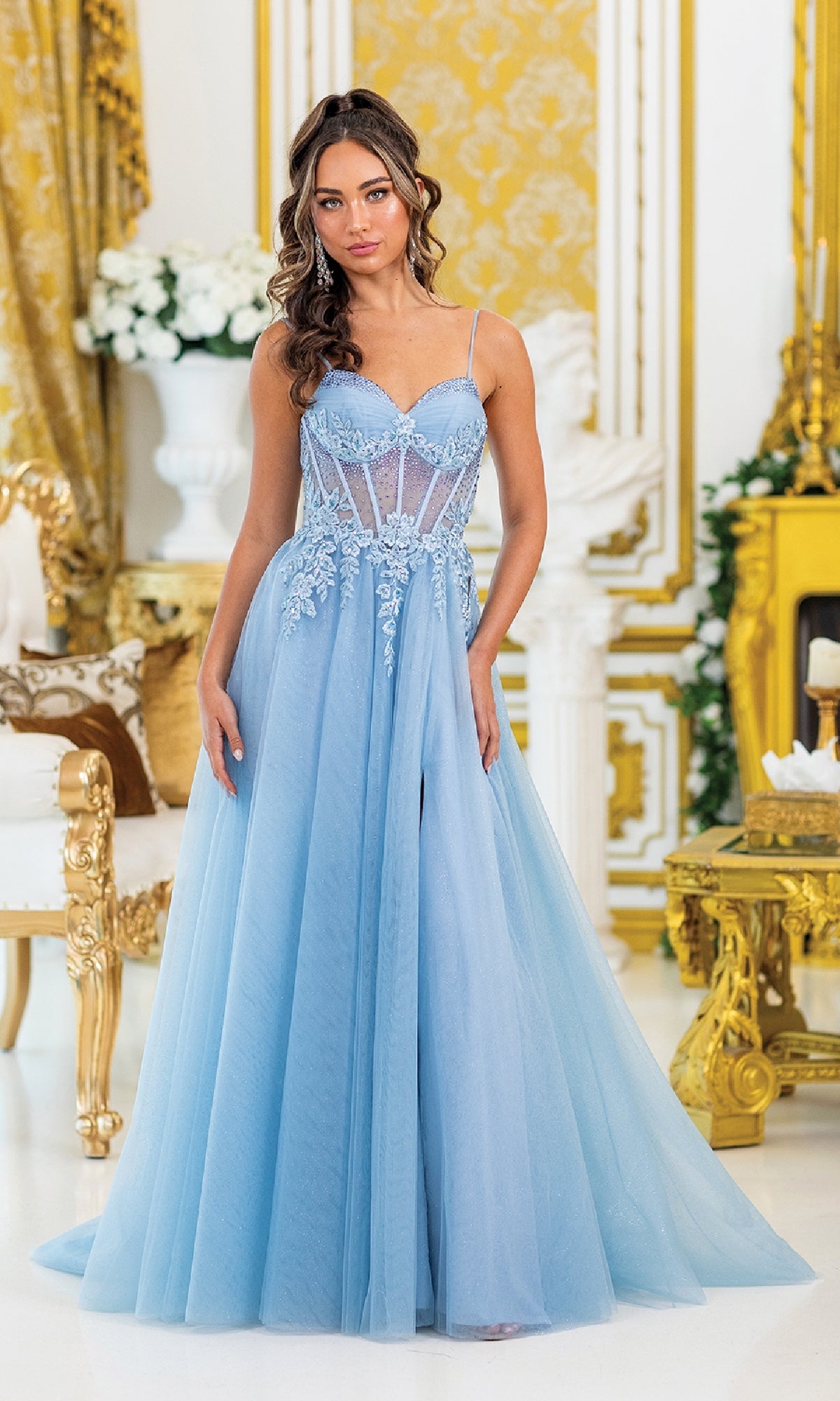 A woman stands indoors in the Corset Prom Ball Gown with Slit: Dancing Queen 4529, a light blue lace embroidered tulle dress with sheer details. The elegant room is decorated with gold and white accents, flowers, and classical furniture.