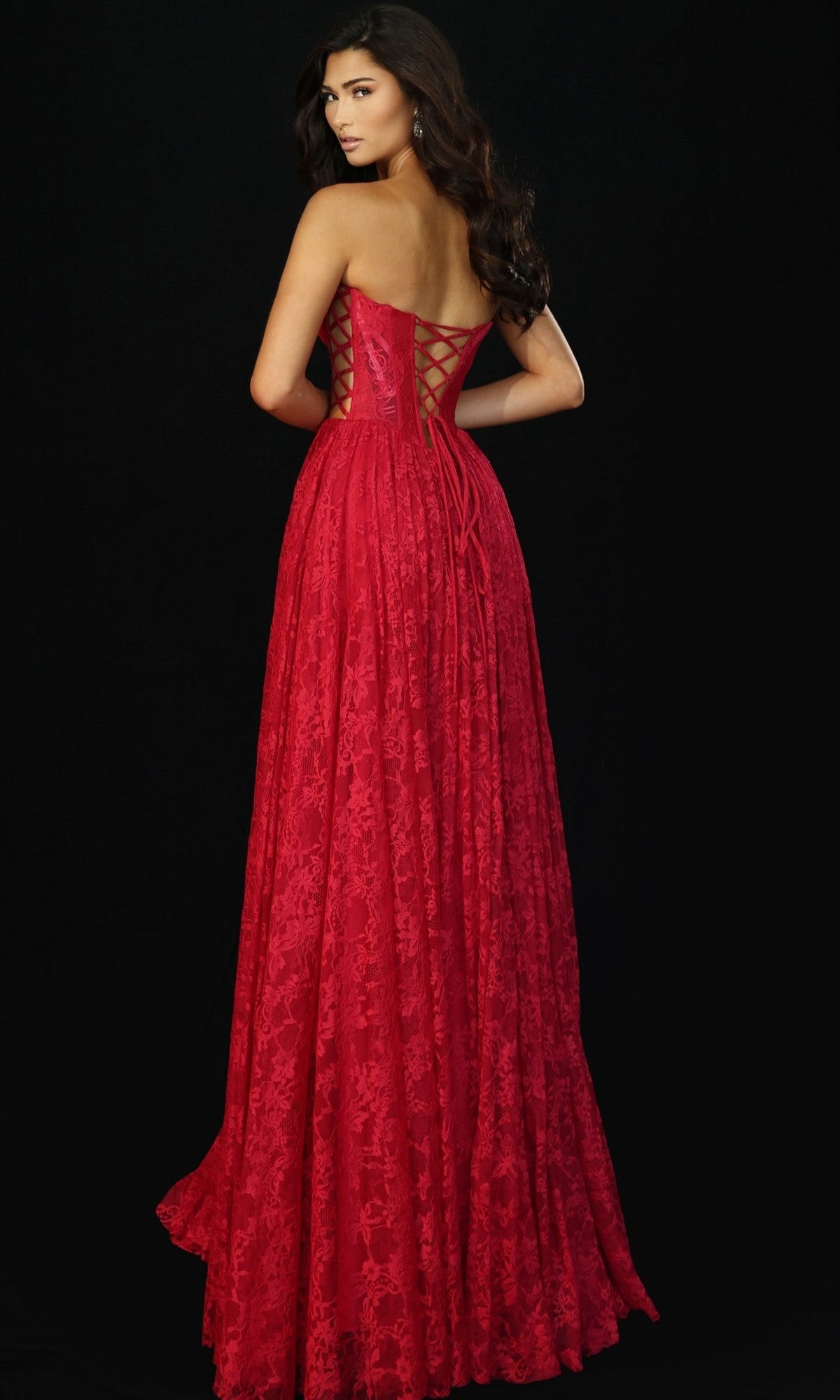 Woman models the Jovani 45372 long prom dress, a strapless gown with a fitted corset bodice, floral lace pattern, and lace-up back. She poses against a black background, glancing over her shoulder.