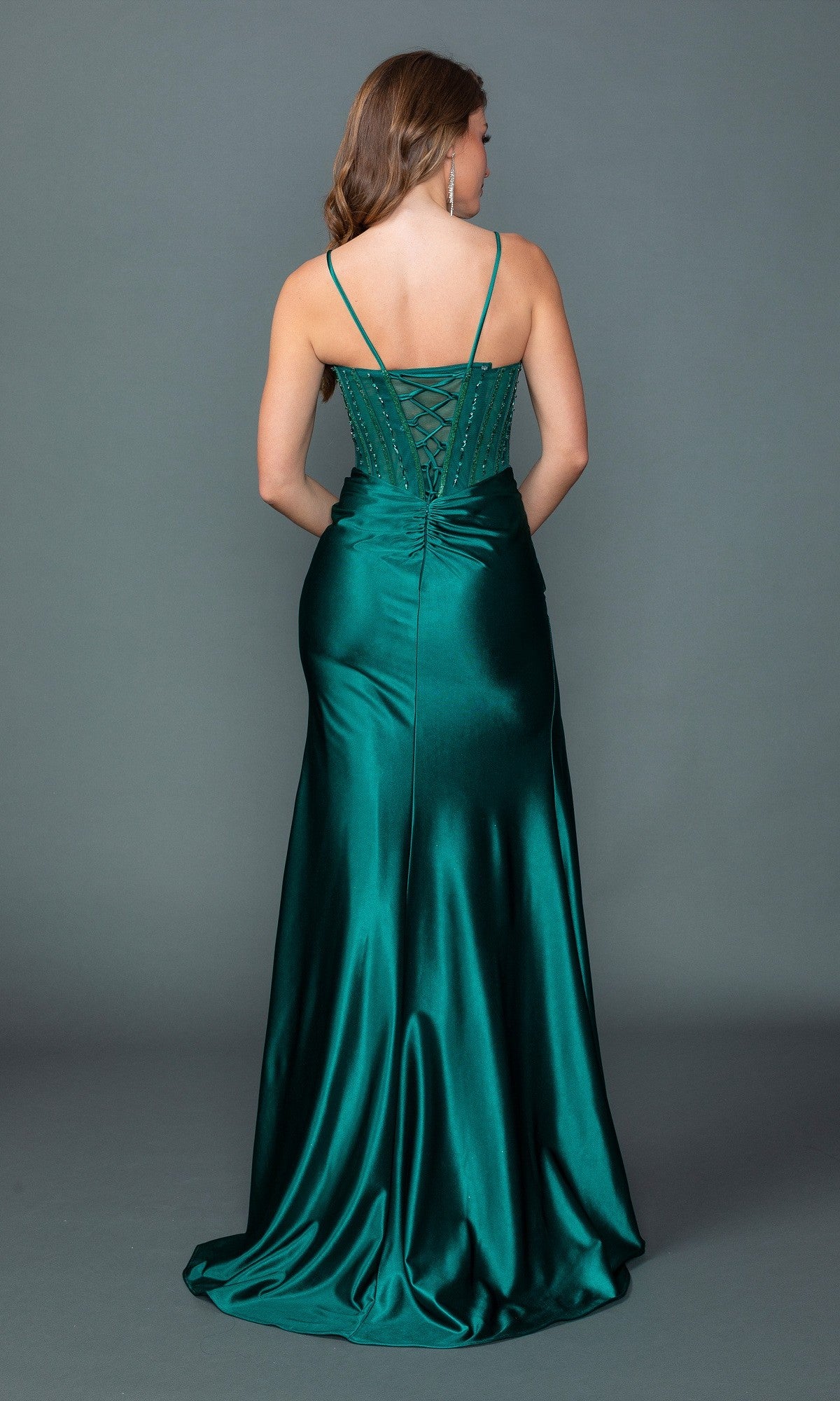 A woman faces away, wearing the Long Prom Dress: Dancing Queen 4561 in emerald green, featuring a high leg slit and lace-up back detail, set against a plain gray background.