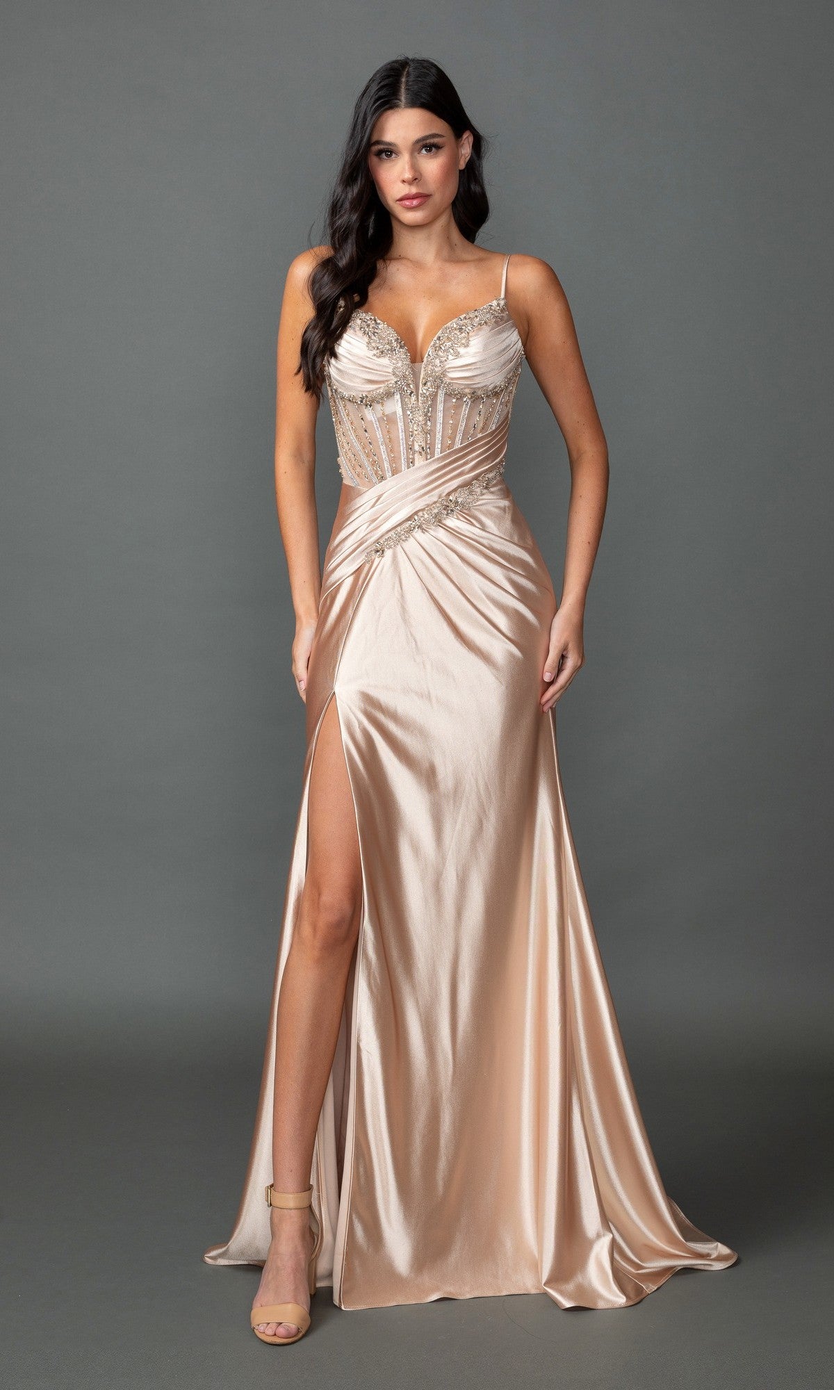A woman wears the Dancing Queen 4561 long prom dress in champagne, featuring a shiny finish, sheer corset bodice, spaghetti straps, and a high leg slit, standing against a plain gray background.