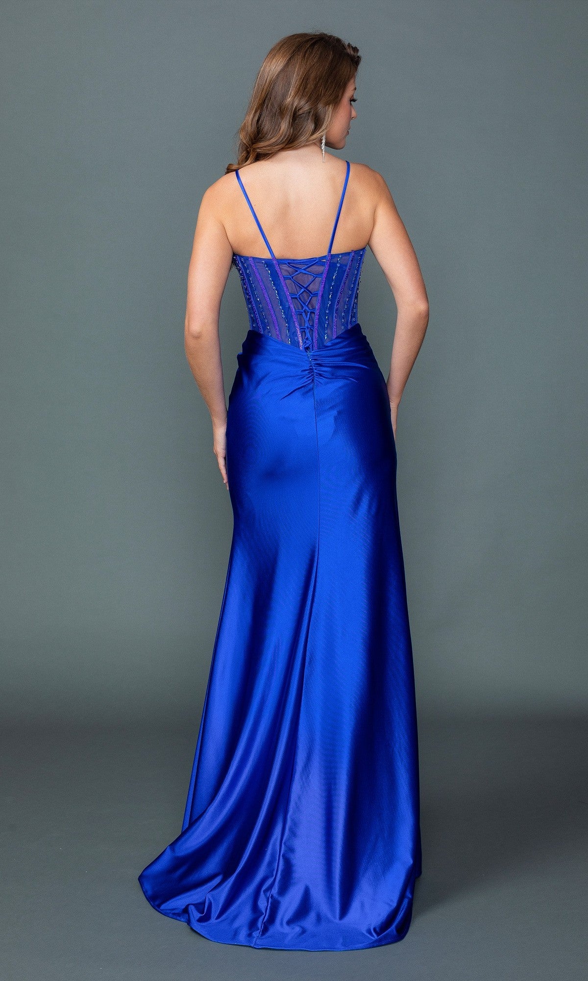 Wearing the Dancing Queen 4561 long prom dress in royal blue satin with spaghetti straps, lace-up back, and high slit, a woman stands facing away from the camera against a plain gray background.