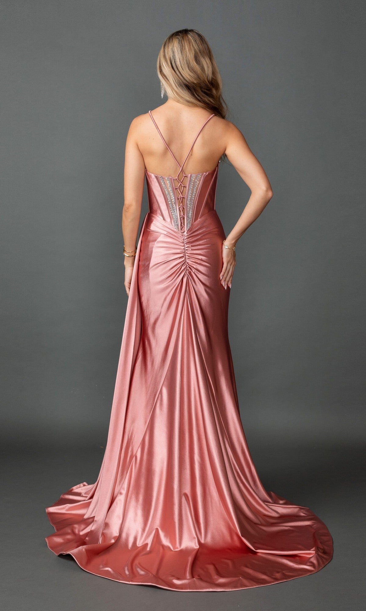 A woman is seen from behind wearing the Long Prom Dress: Dancing Queen 4581—a shiny, floor-length rose-colored gown with thin straps, a ruched back detail, and a sleek satin sheath skirt—set against a plain gray background.